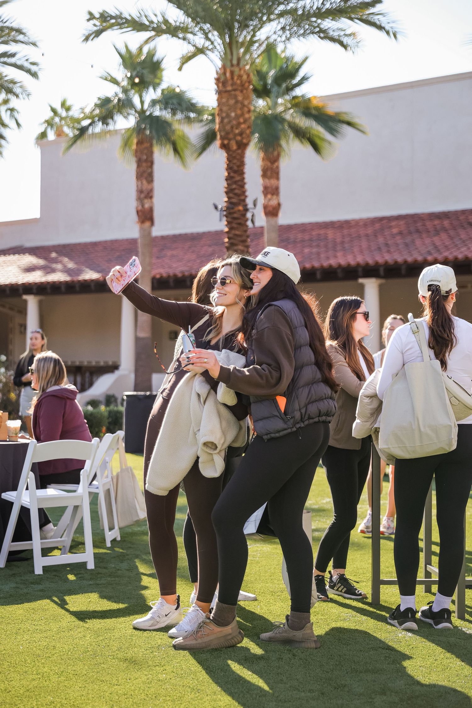 Two women take a selfie on a sunny lawn at an outdoor event with palm trees and white chairs.
