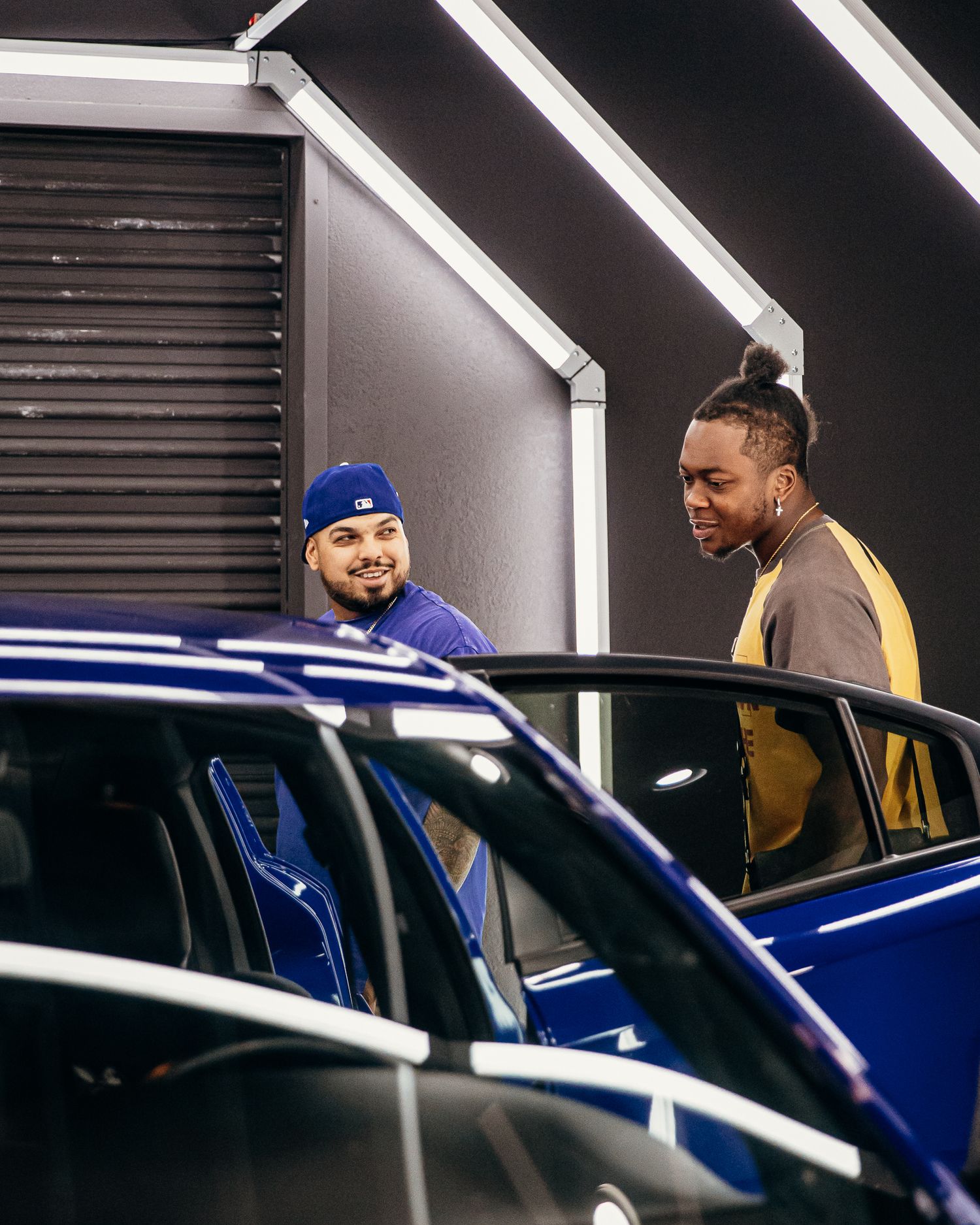 Two men examining a blue luxury car in a modern showroom, one wearing a blue cap and the other with a bun hairstyle.