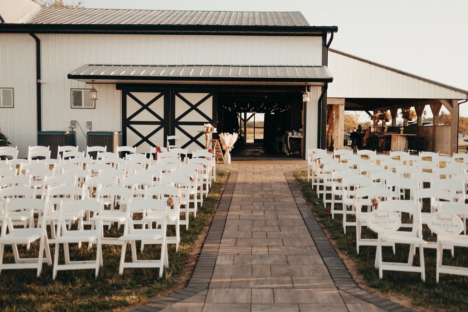 Rows of white chairs line a brick aisle leading to a rustic barn wedding venue at sunset.