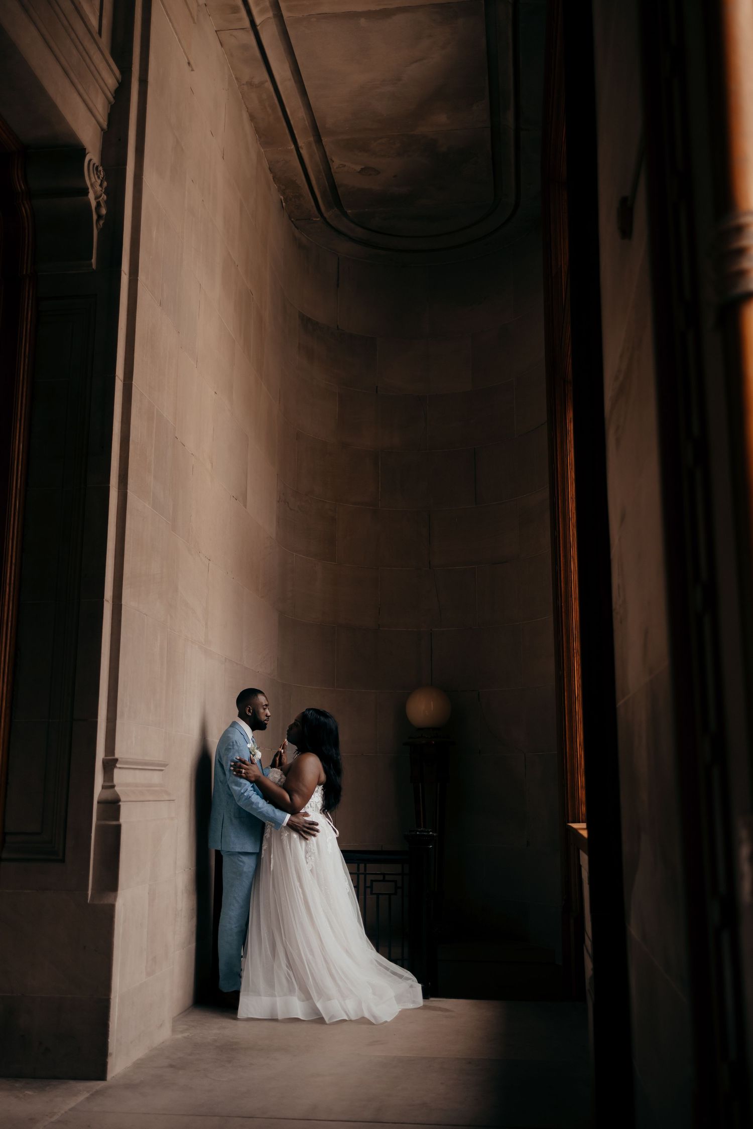 A bride and groom share a romantic embrace in a grand architectural hallway, bathed in soft dramatic light.