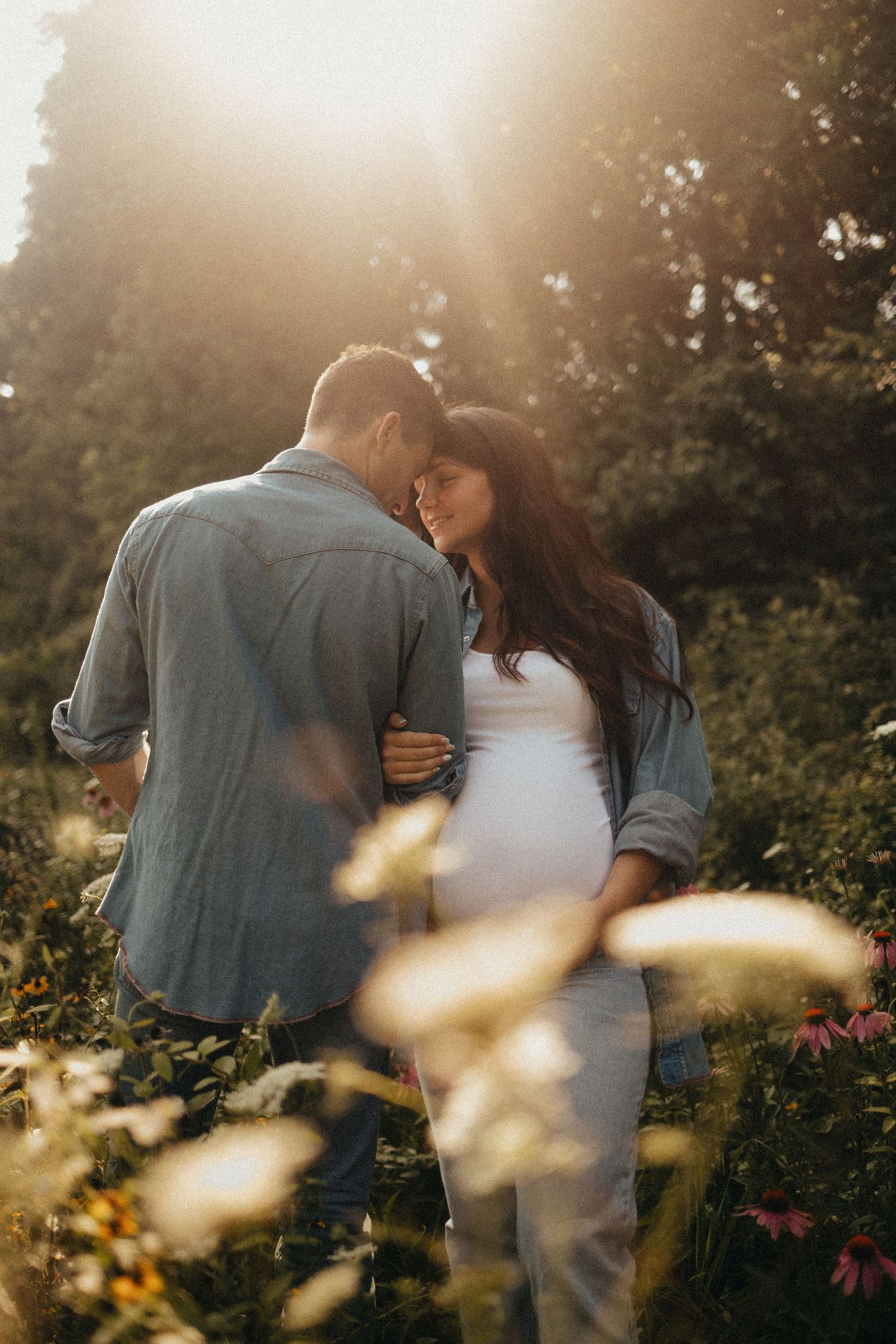Pregnant couple embracing in a sunlit wildflower garden during golden hour.