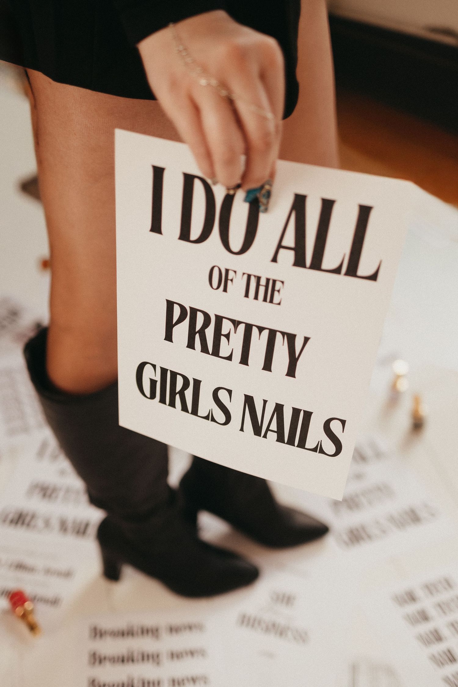 Person holding a sign reading I Do All of the Pretty Girls Nails while wearing black boots.