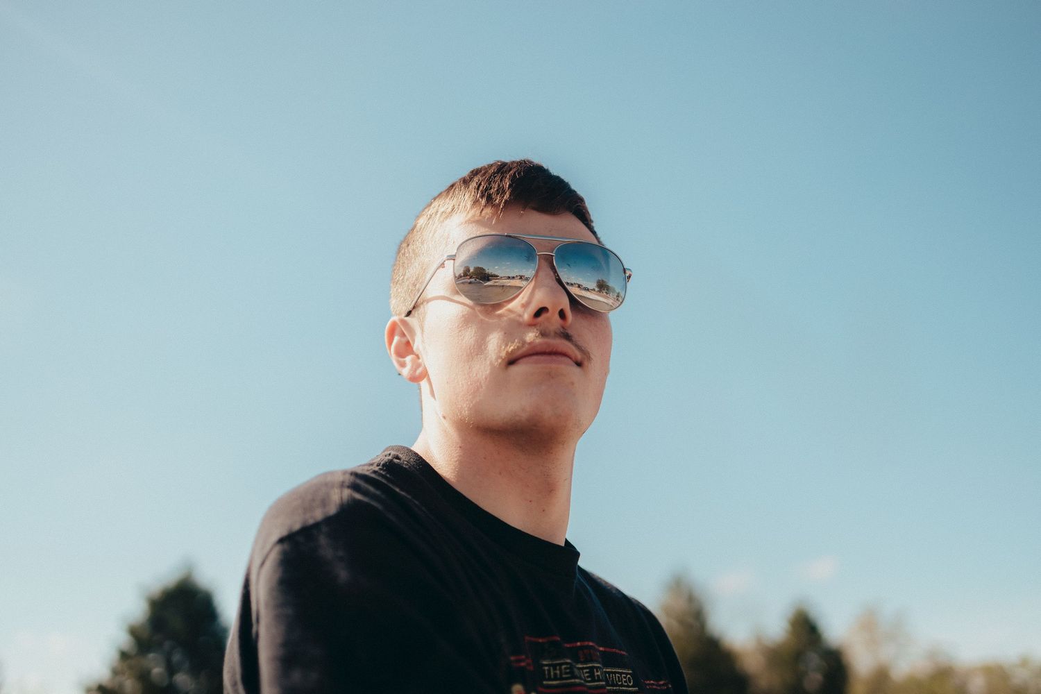 Young man wearing reflective aviator sunglasses looking up against a clear blue sky outdoors.