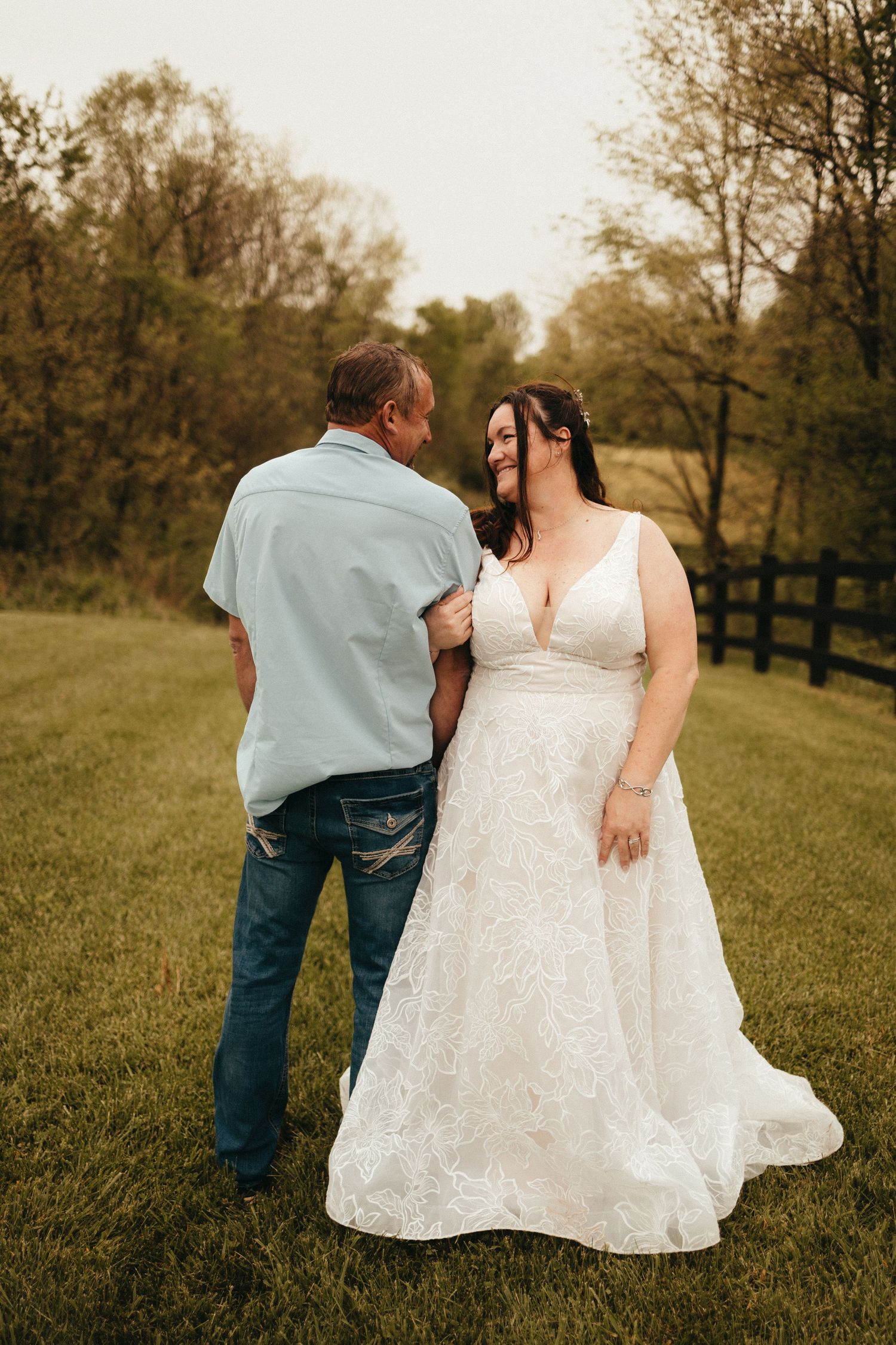 Bride in lace wedding gown walks with man in blue shirt and jeans on green lawn with wooden fence.