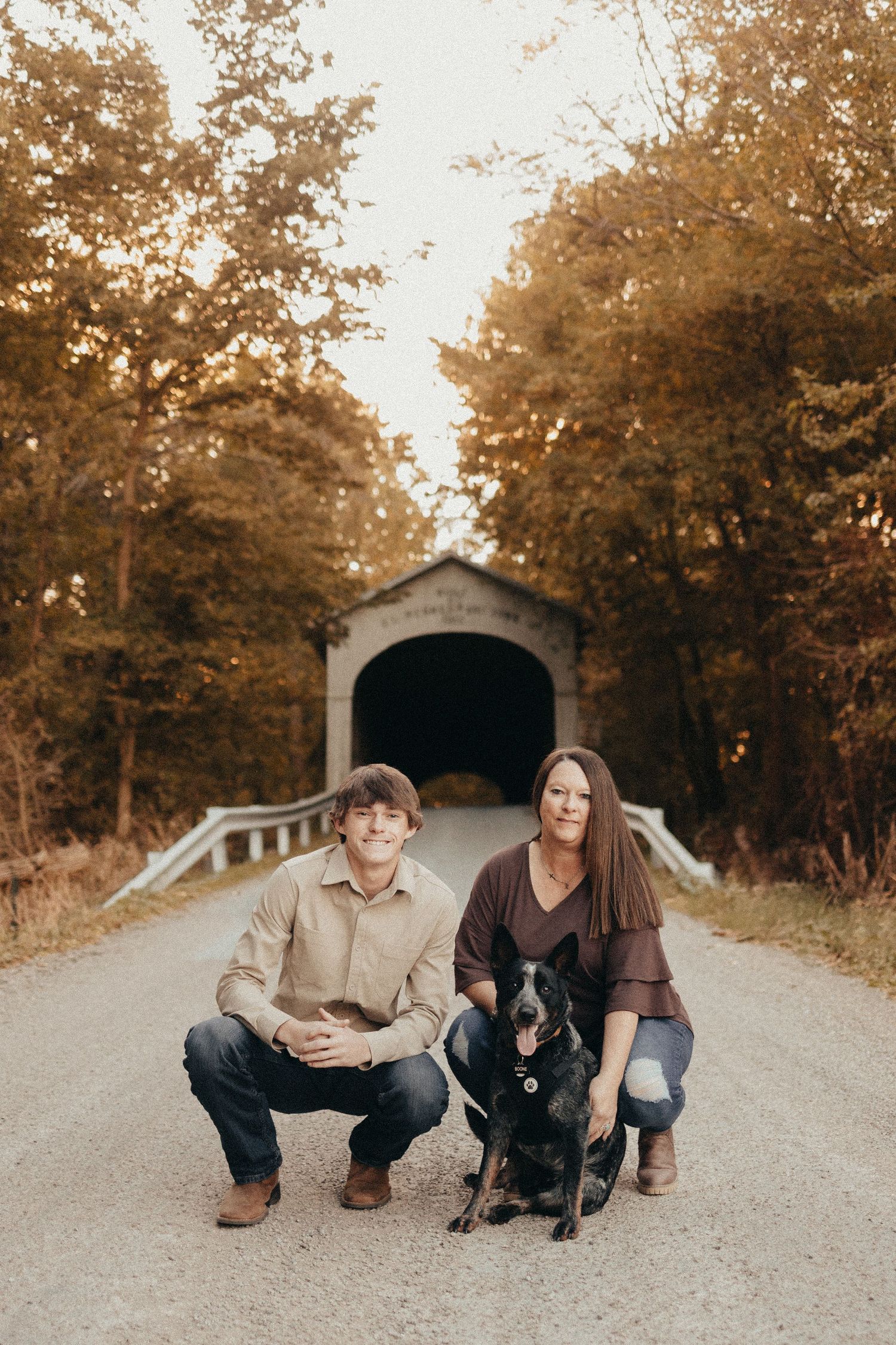 A couple crouches with their black dog on a gravel road in front of a historic covered bridge surrounded by fall foliage.