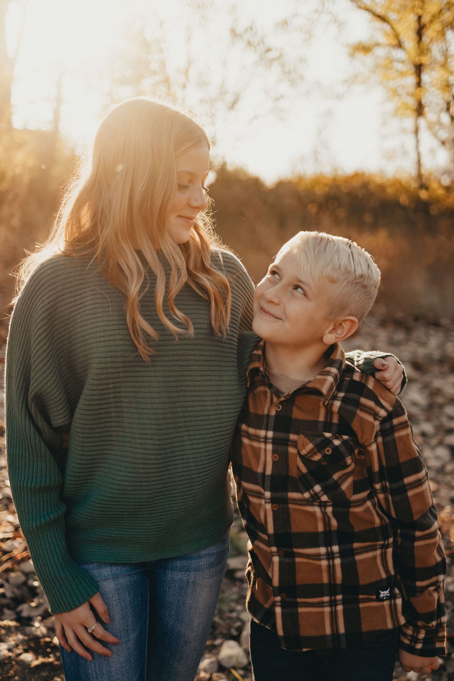 A blonde woman in a green sweater and a short-haired boy in plaid share a warm moment in golden sunset light.
