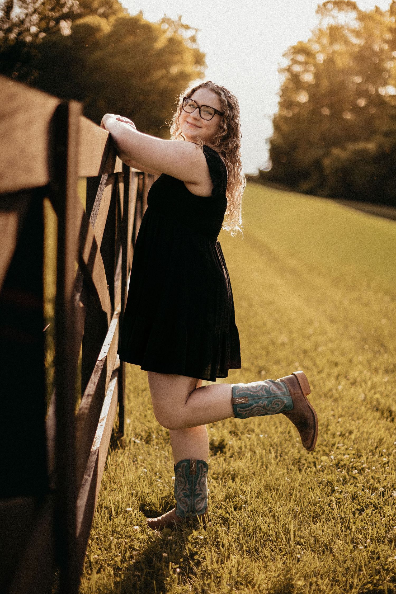Young woman in black dress and cowboy boots leaning on a wooden fence at golden hour.
