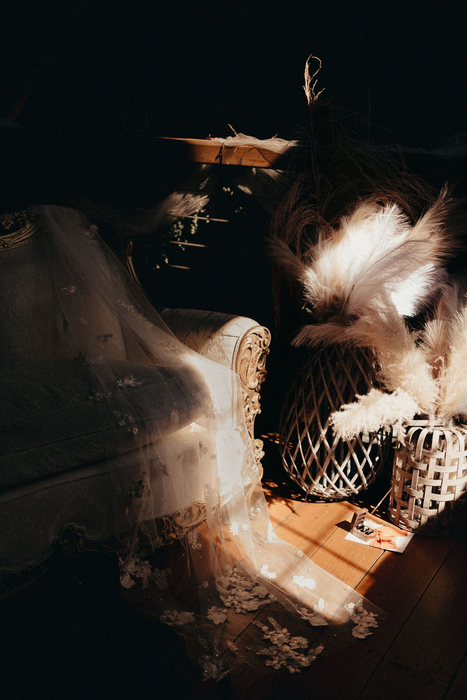 Elegant vintage wedding setup with ornate chair, white feathers, and floral veil in dramatic moody lighting.