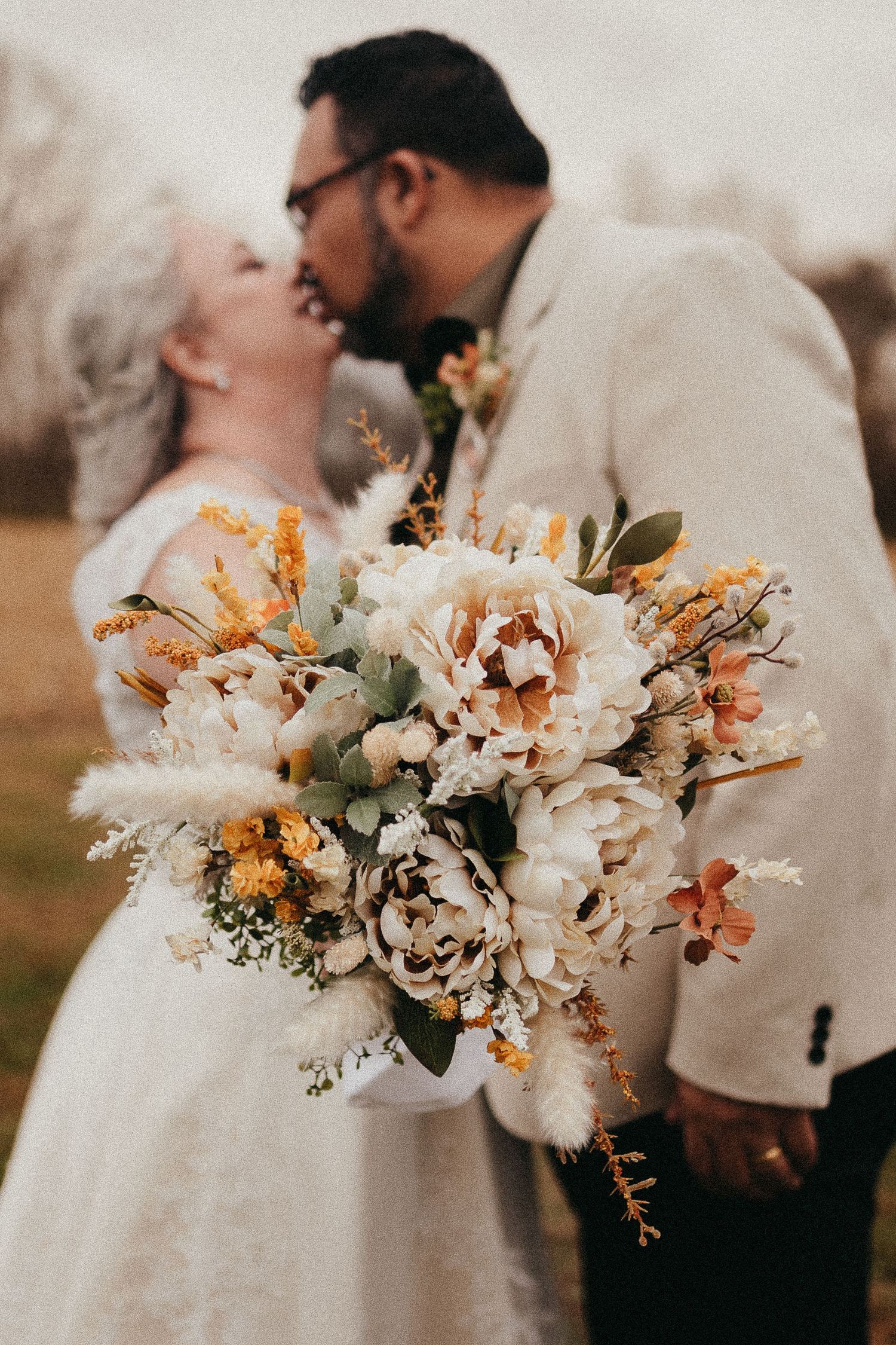 Couple kissing behind stunning boho bridal bouquet of cream peonies and yellow wildflowers.