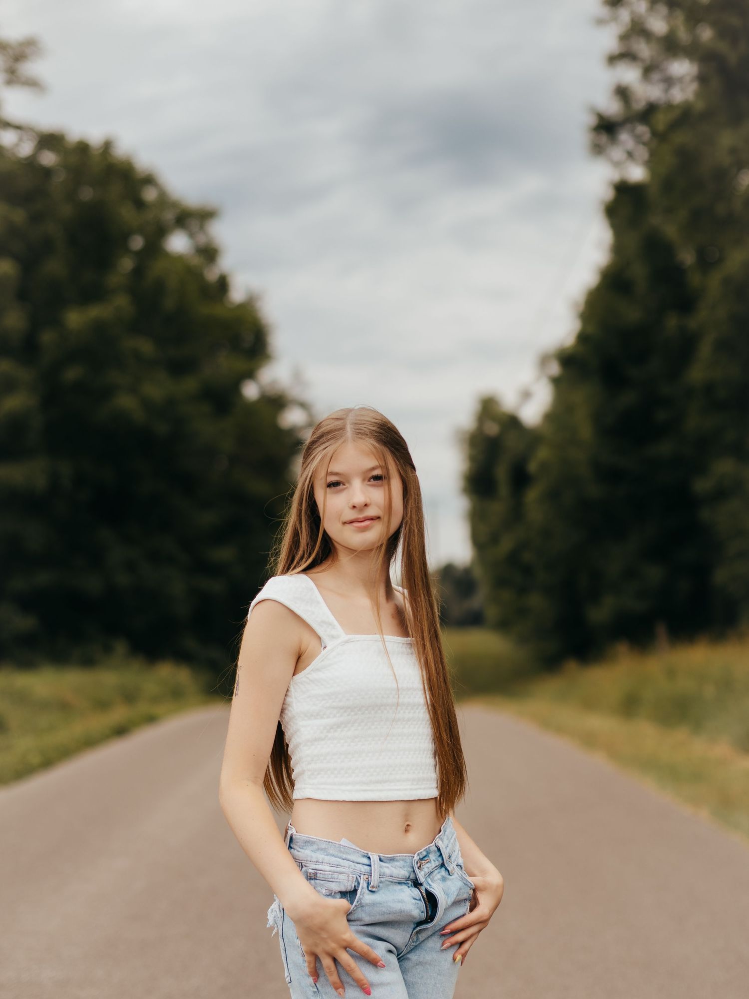 Young girl with long hair in white crop top and jeans posing on a tree-lined road under cloudy skies.