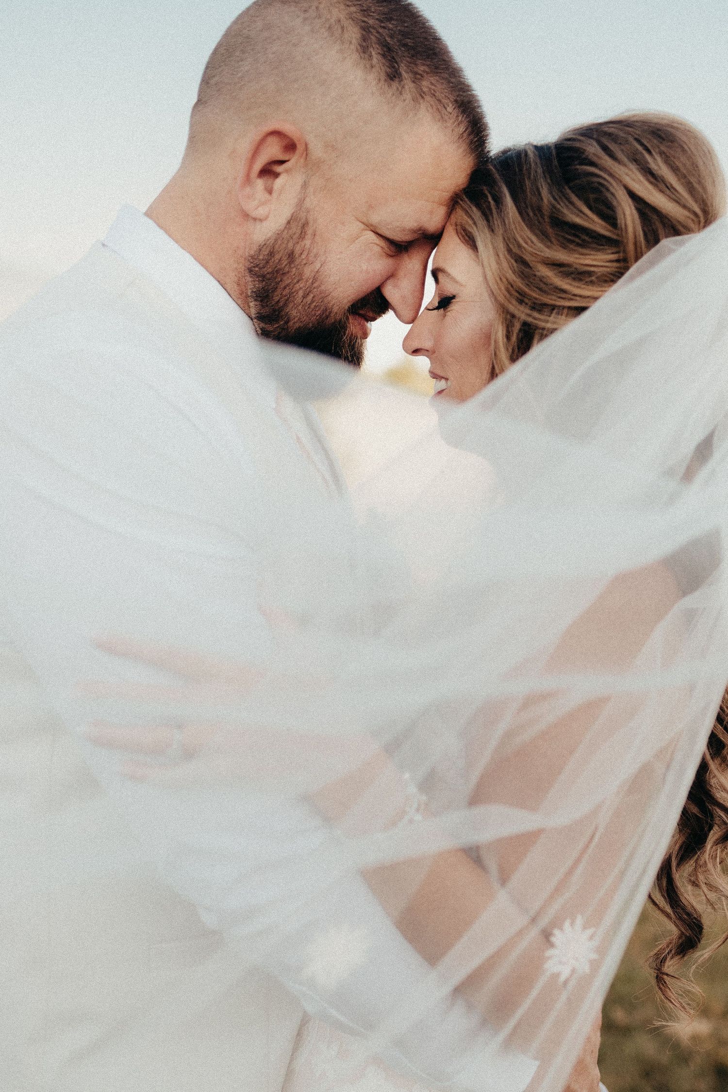 Romantic bride and groom touching foreheads under a flowing white veil during an outdoor wedding photo shoot.