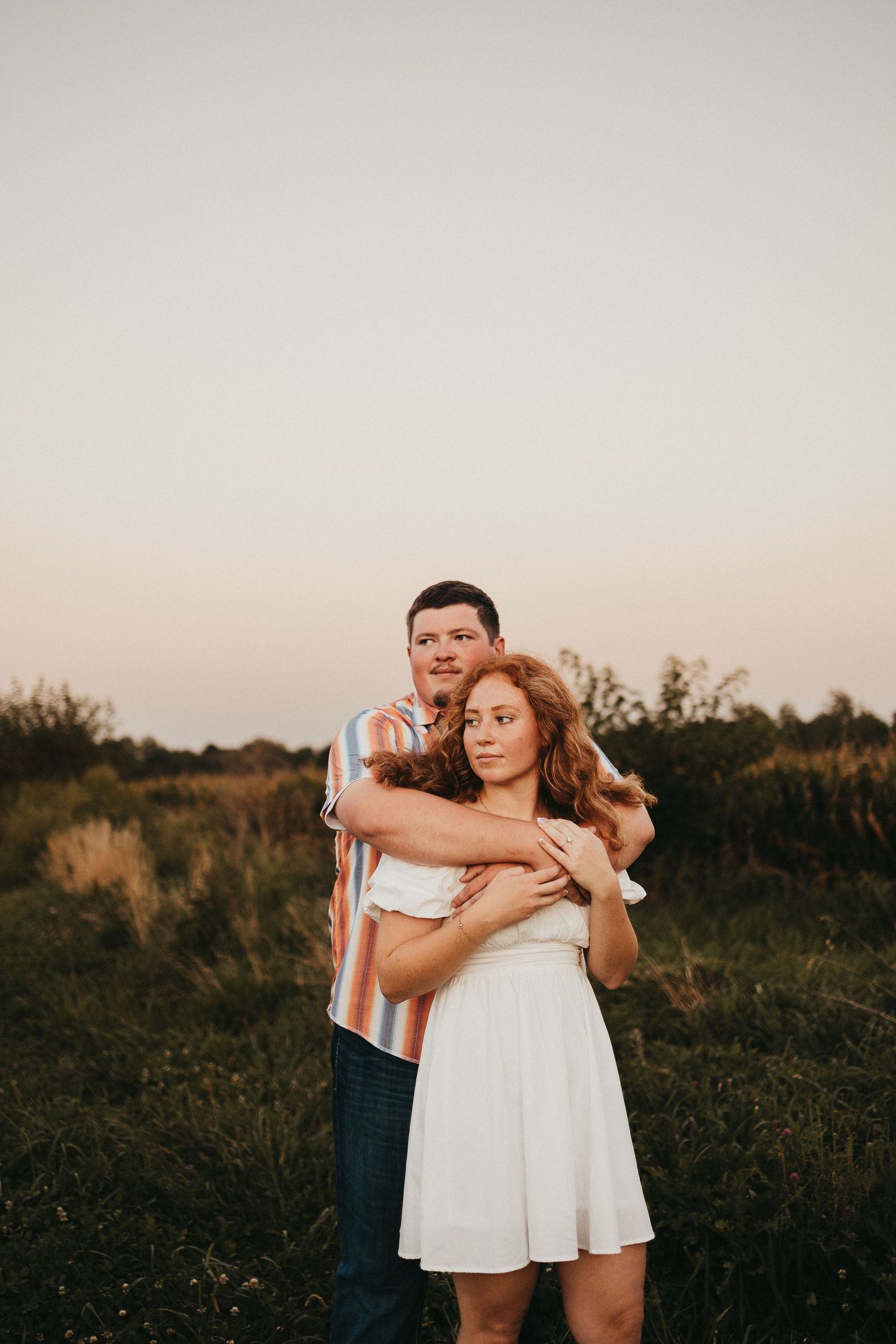 Couple embracing in a golden hour field, woman in white dress, man in striped shirt, romantic outdoor portrait.