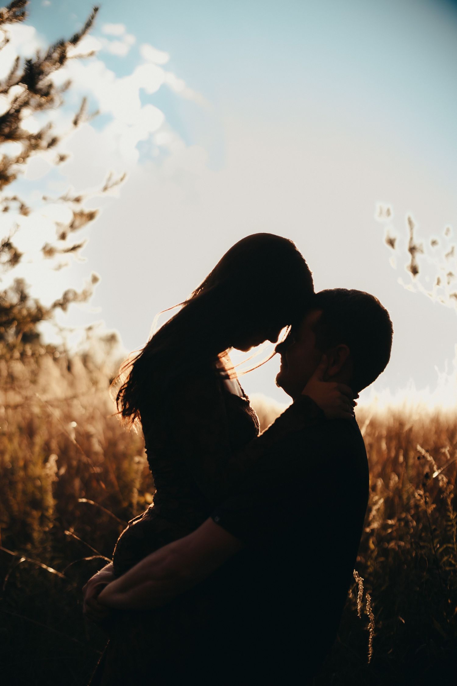 Silhouette of a couple embracing forehead to forehead in a golden field at sunset with soft backlight and tall grass.