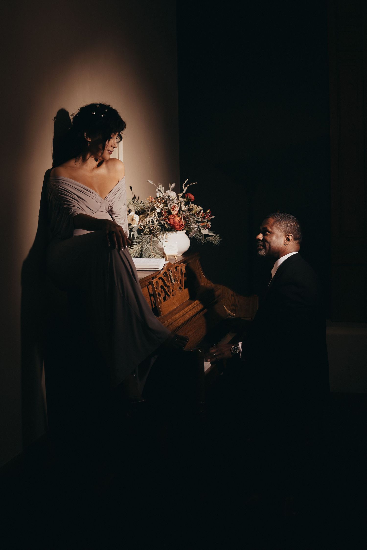 A man in a suit kneels before a woman in a gray gown beside a piano with floral arrangement in dramatic lighting.