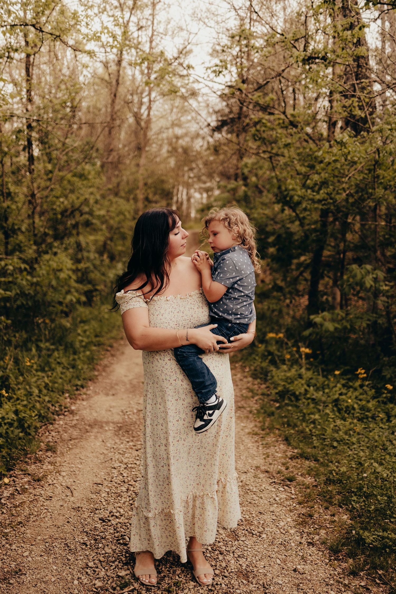 Mother in white dress holding and gazing at her young child on a wooded forest path.