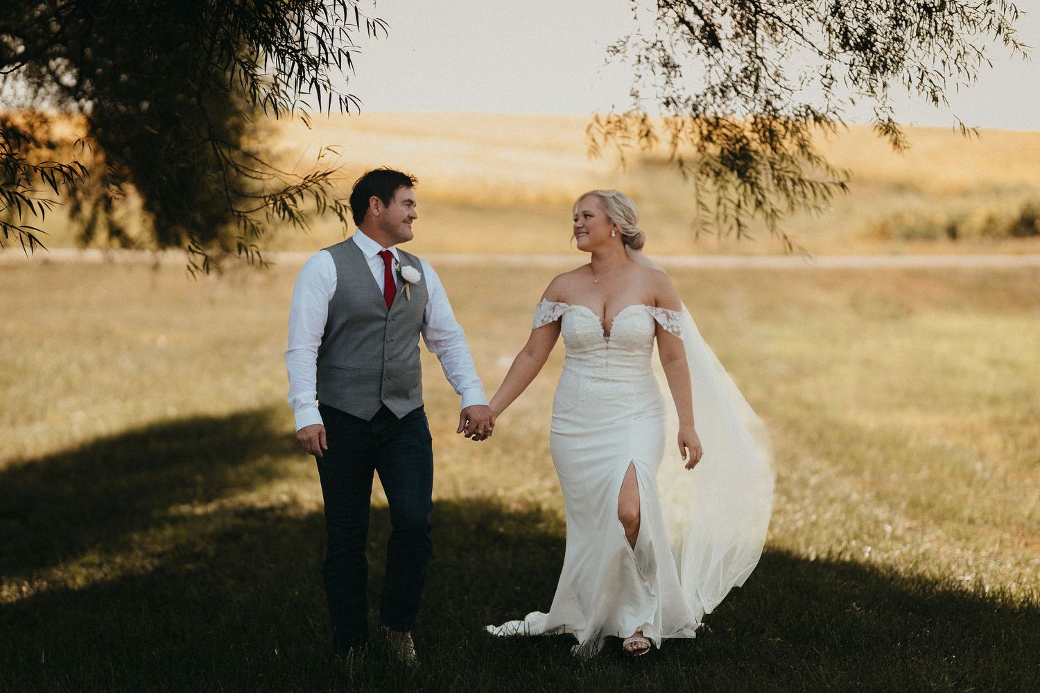 Bride and groom walking hand in hand through golden field at sunset, bride in off-shoulder gown with flowing skirt.