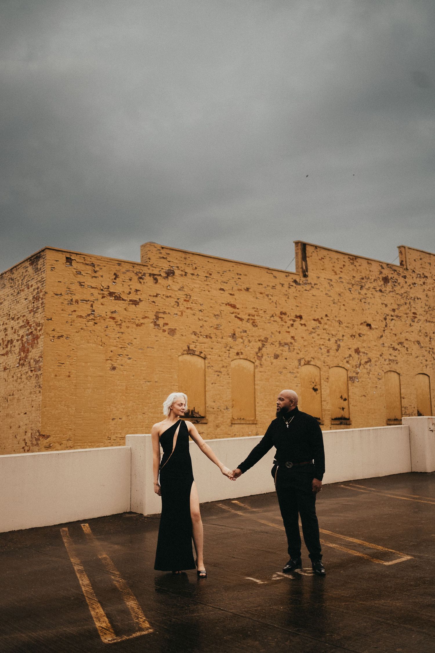 Couple in elegant black attire holding hands on a rooftop parking lot with a yellow brick building and cloudy sky.
