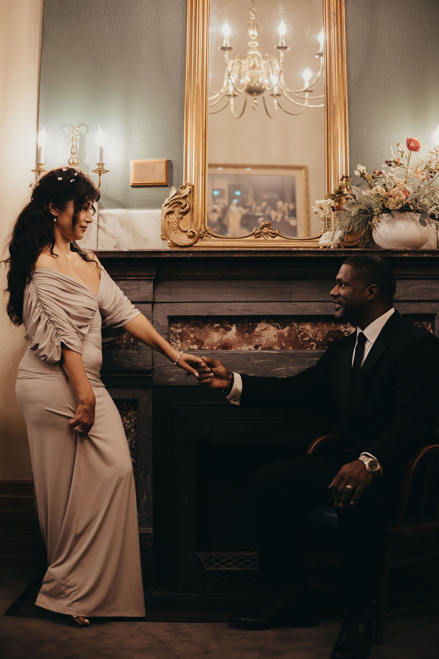 Elegant couple in formal attire sharing a moment by an ornate fireplace with gold mirror and chandelier in a luxurious room.