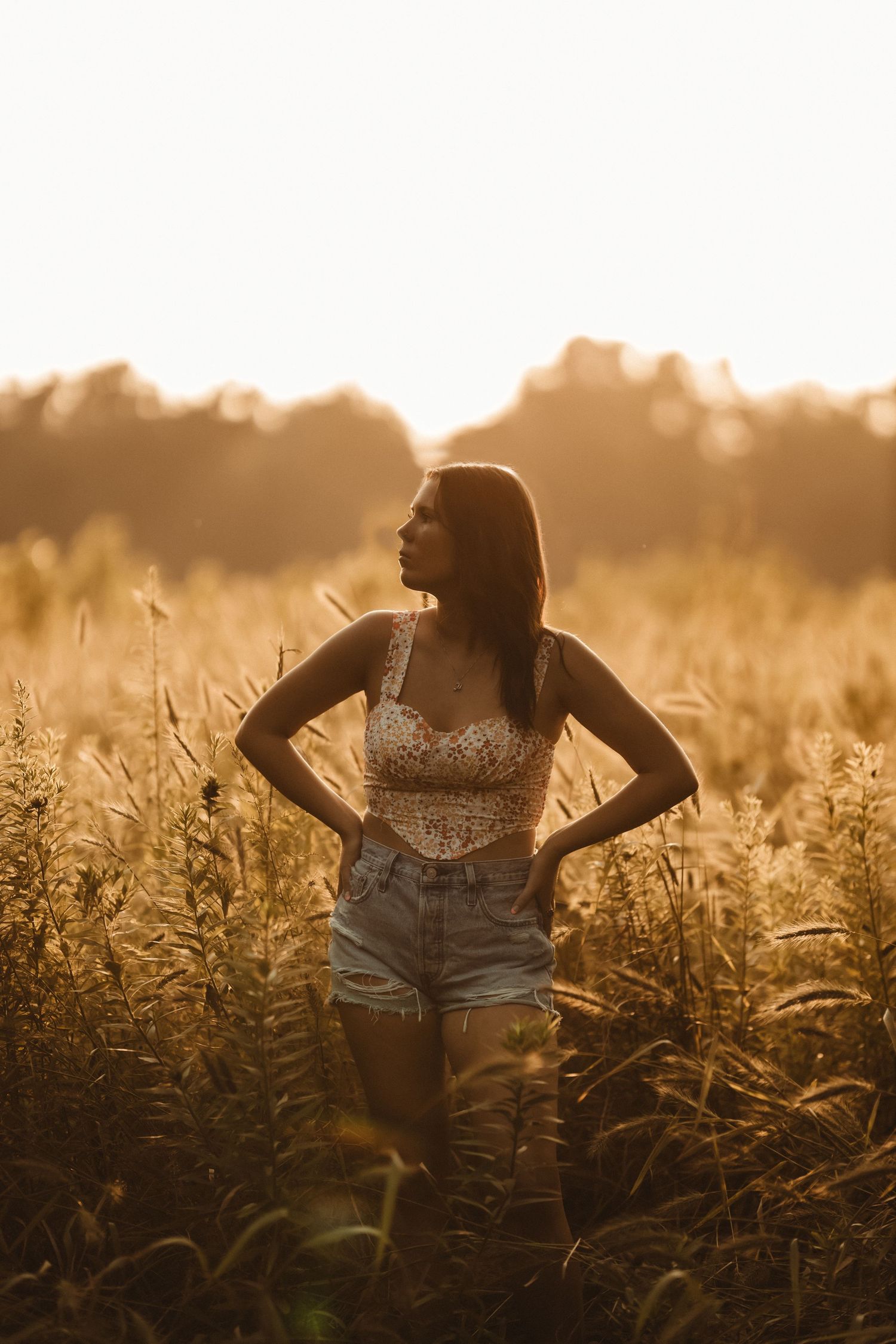 Woman in floral crop top and denim shorts standing in golden wheat field during sunset.