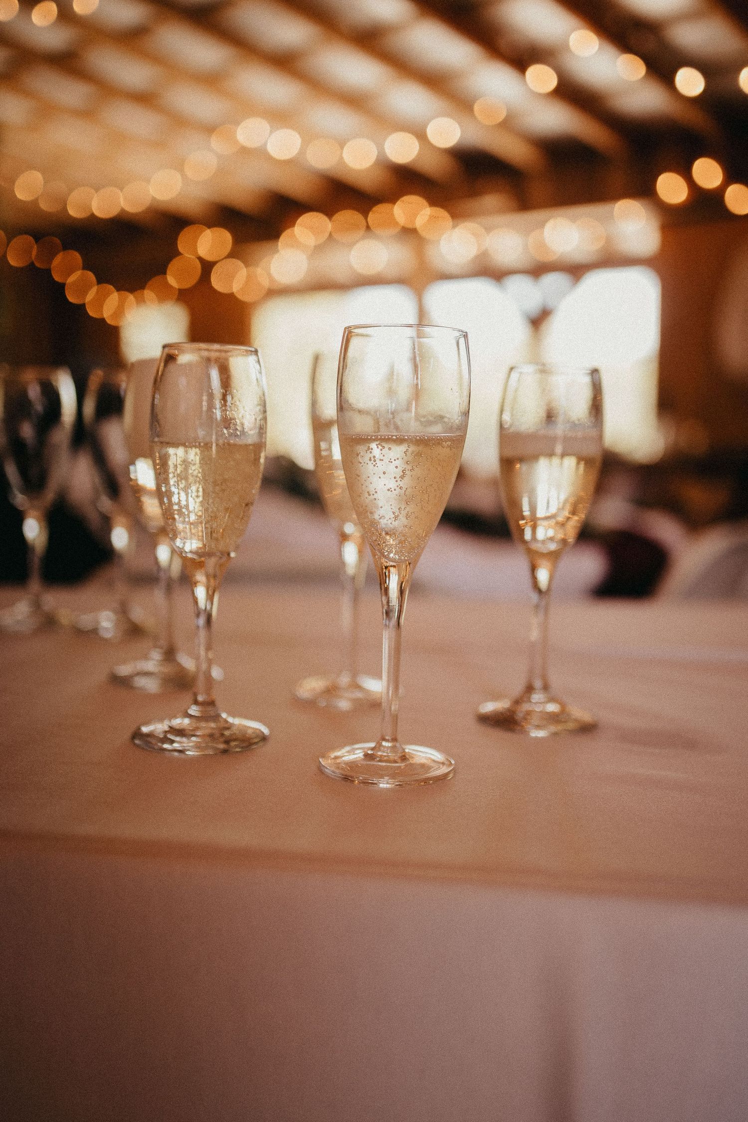 Elegant champagne flutes filled with sparkling wine lined up on white tablecloth with warm bokeh lights behind.