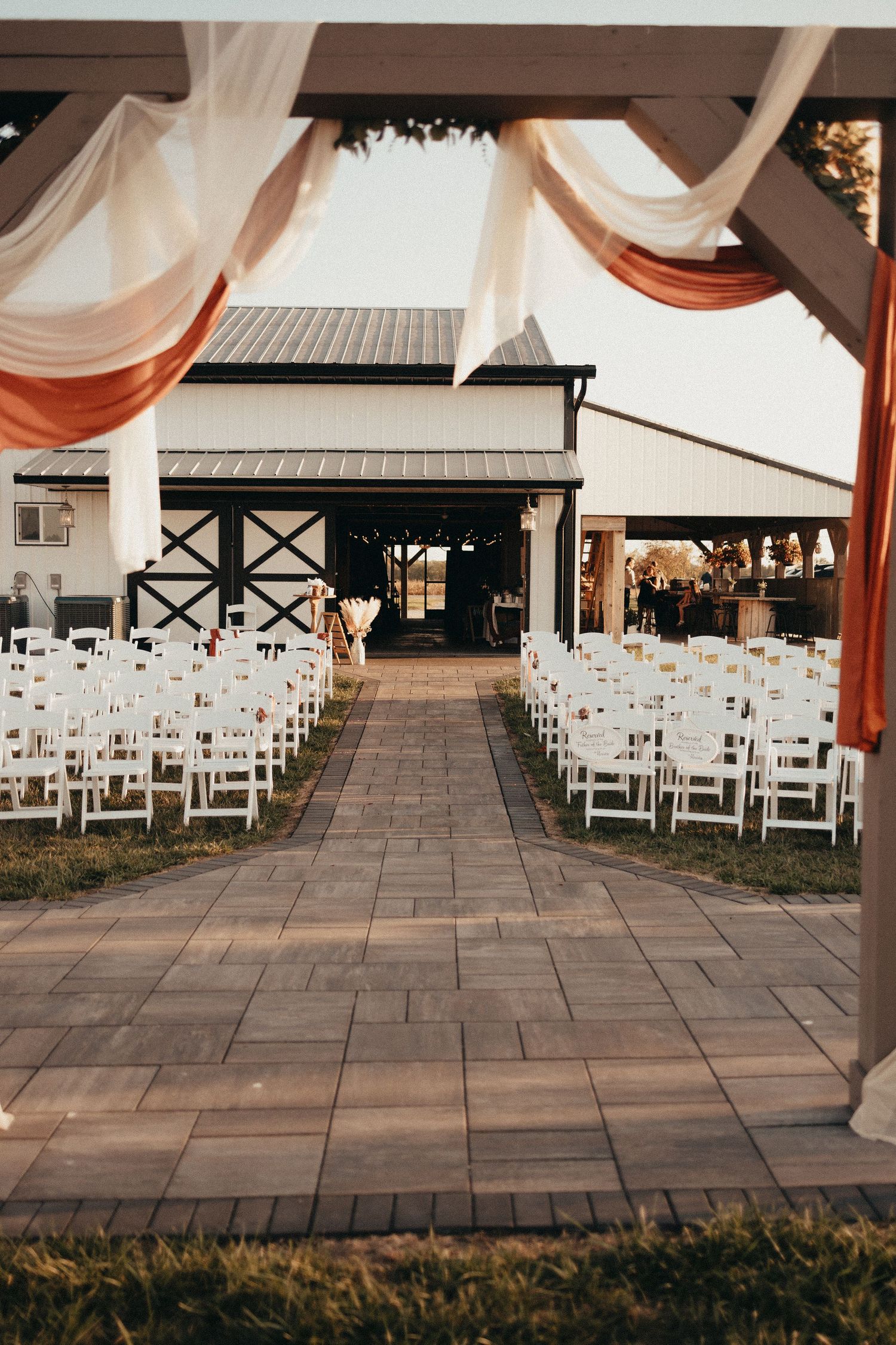 Outdoor barn wedding ceremony aisle with white chairs and terracotta draped pergola at sunset.
