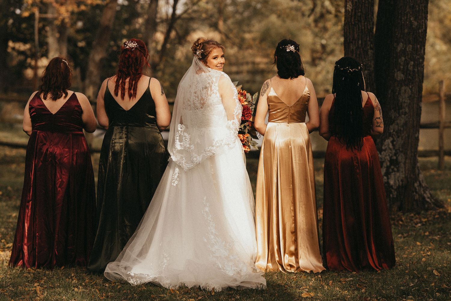 Bride and four bridesmaids in jewel-tone velvet gowns pose with their backs to the camera in an autumn outdoor setting.