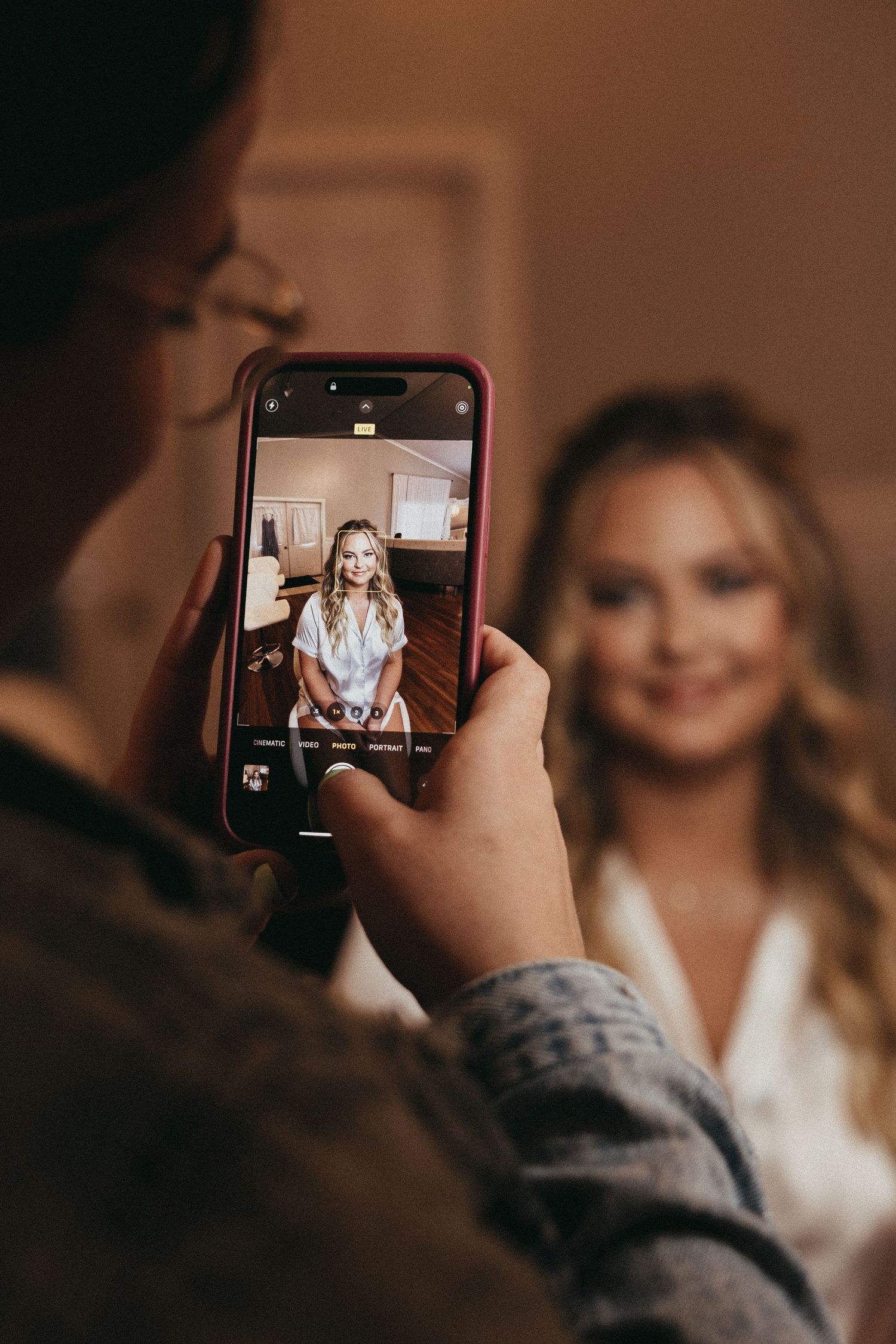 Person photographs smiling blonde woman in white robe with pink-cased smartphone indoors.