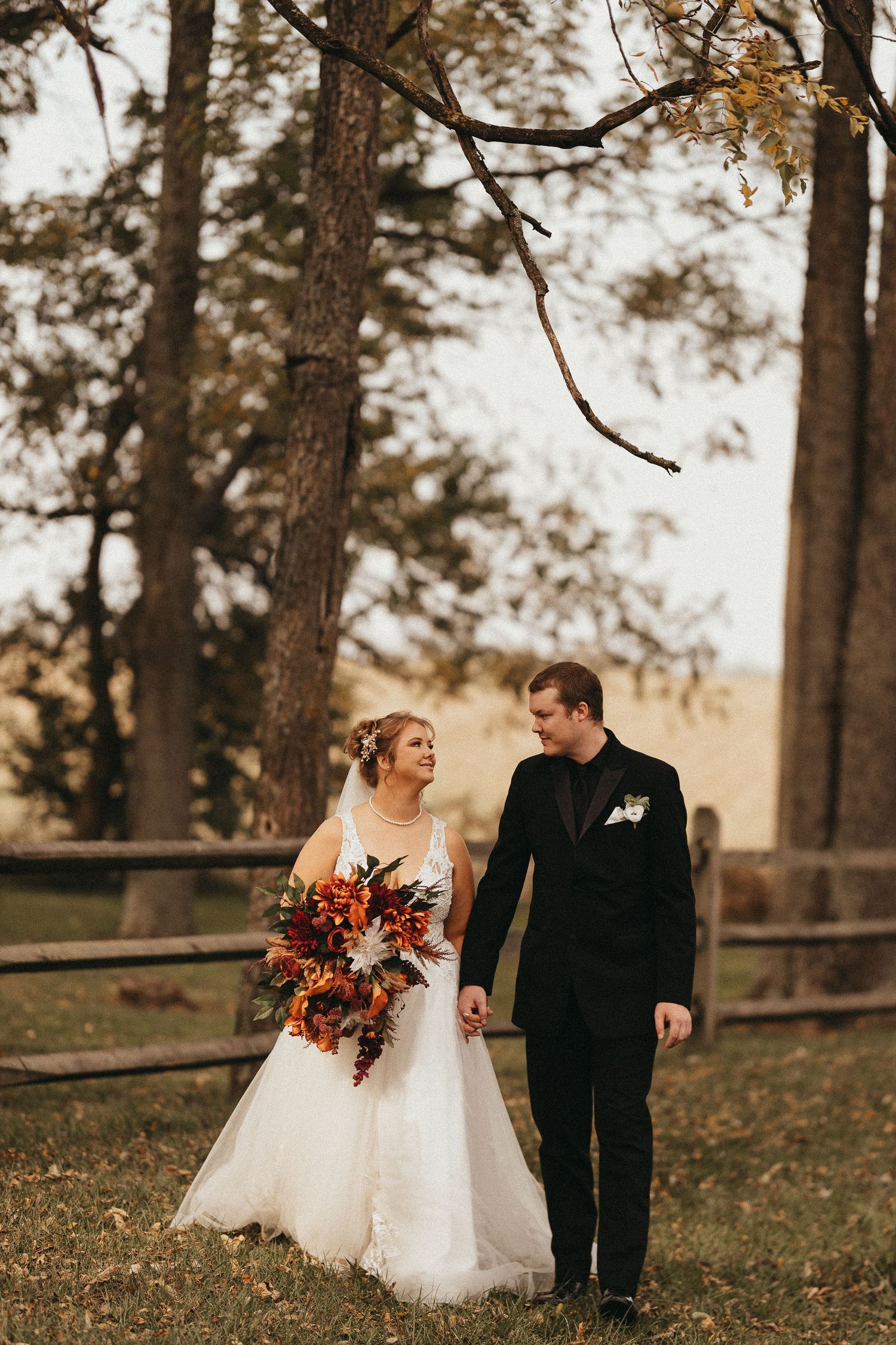 Bride and groom walk hand in hand through an autumn landscape with tall trees and a wooden fence in the background.
