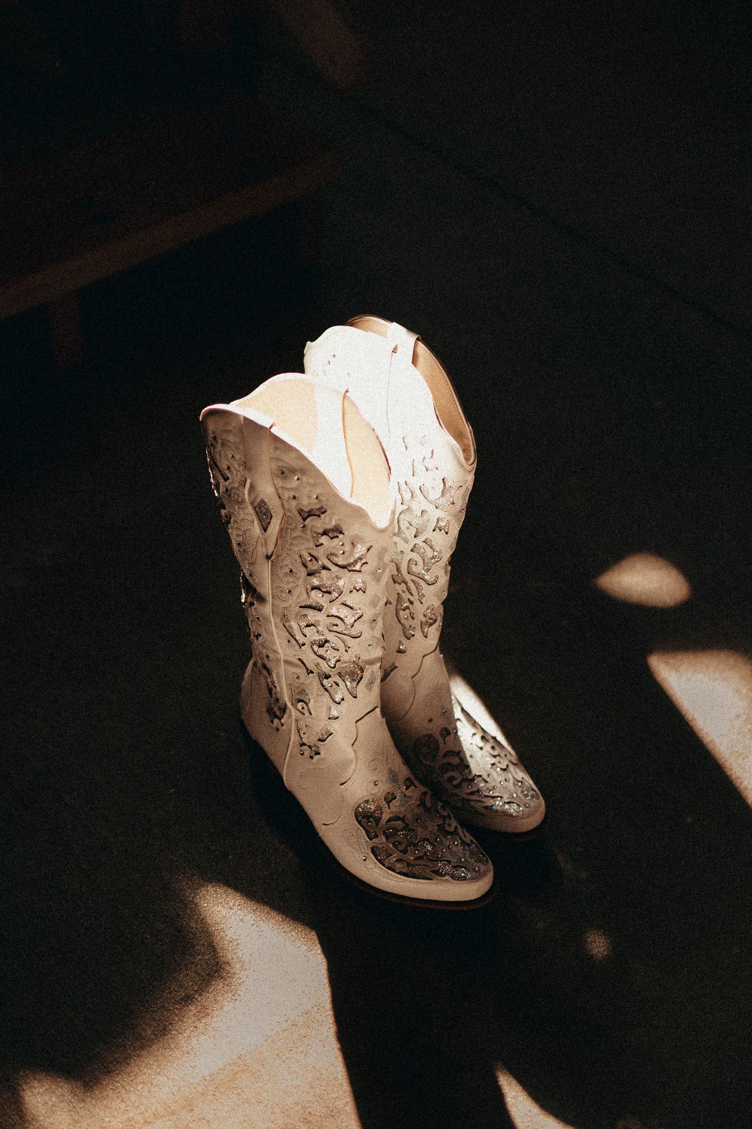 White embroidered cowboy boots with floral details sitting in warm sunlight on dark floor.
