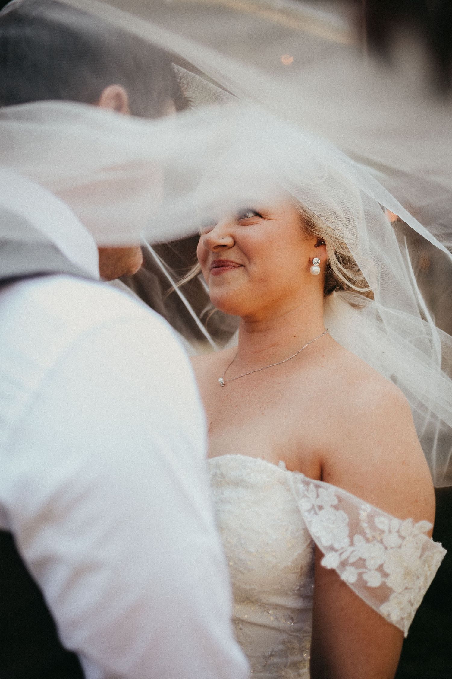 Bride smiling up at groom under flowing white veil wearing off-shoulder lace wedding dress with pearl earrings.