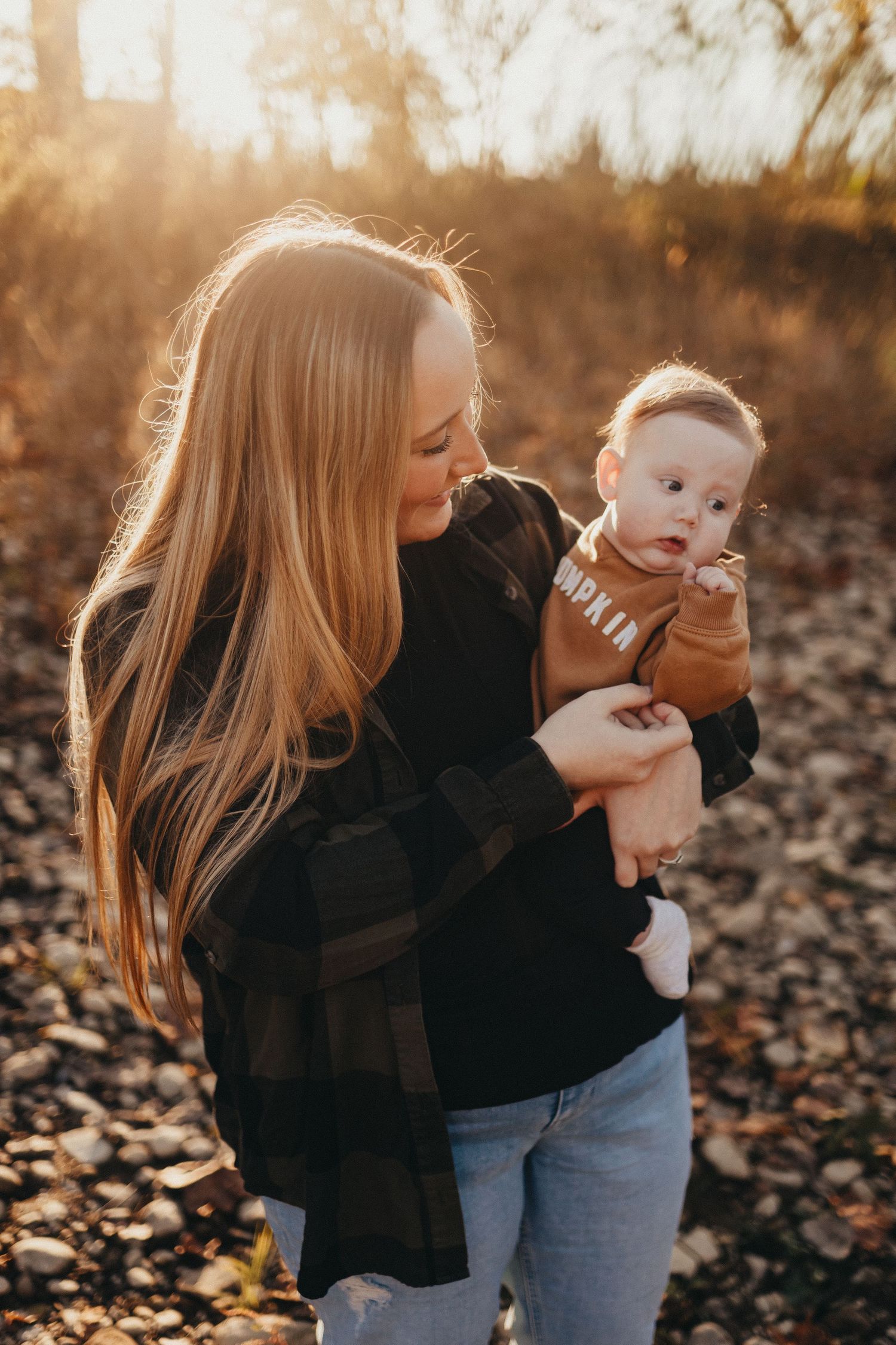 A young mother with long blonde hair tenderly holds her baby outdoors in beautiful warm golden hour sunlight.