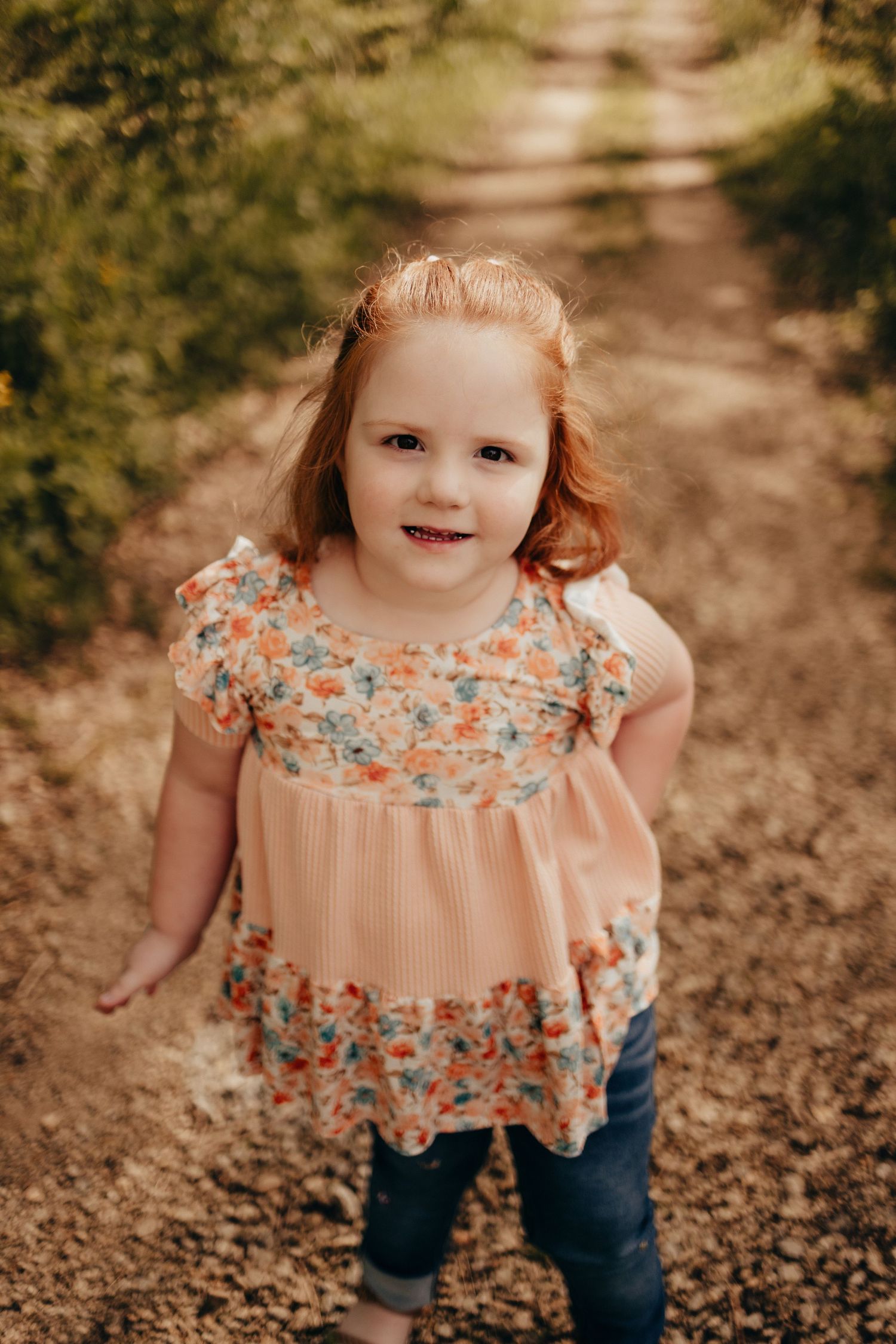 Adorable red-haired little girl in a floral dress smiling on a dirt path outdoors.