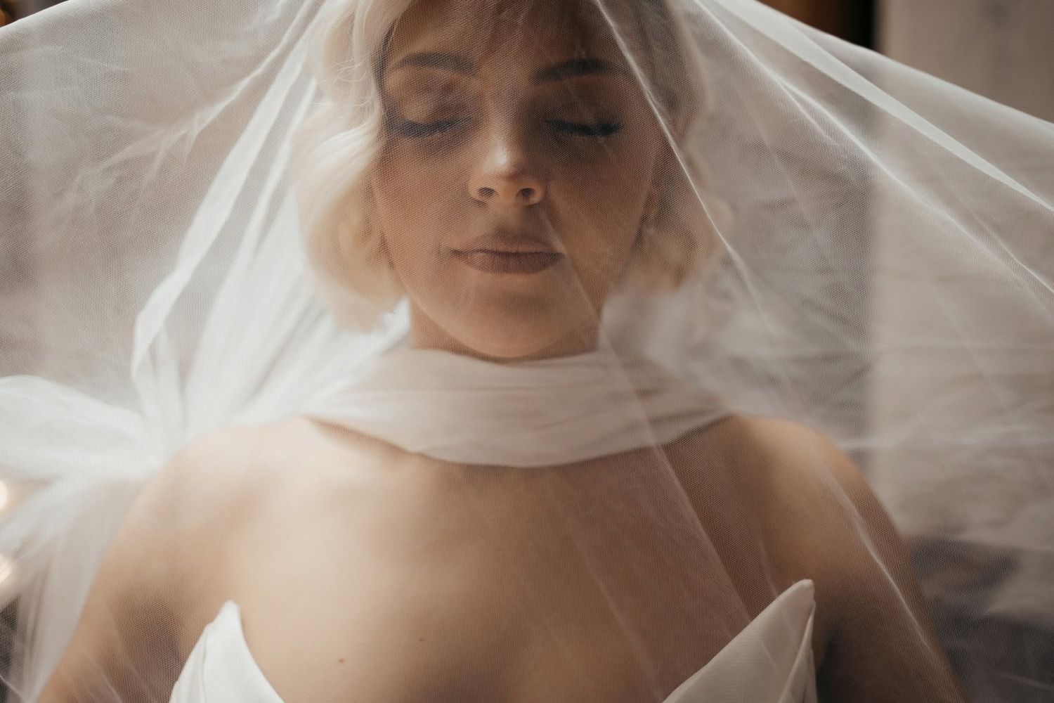 A bride wearing a white veil draped over her head with eyes closed in a serene and elegant wedding portrait.