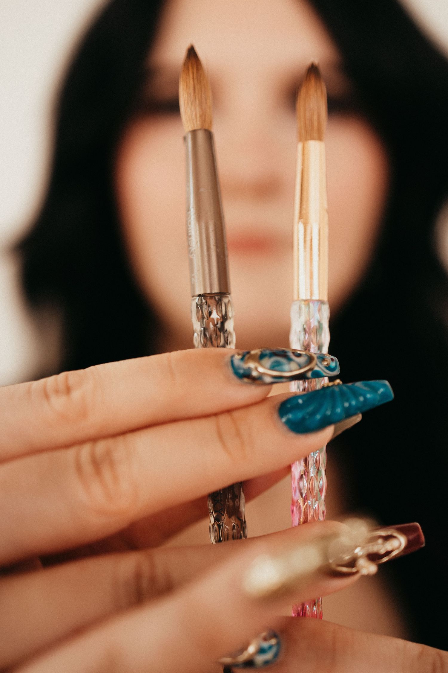 Close-up of elaborately decorated long nails holding two nail art brushes with intricate 3D designs.