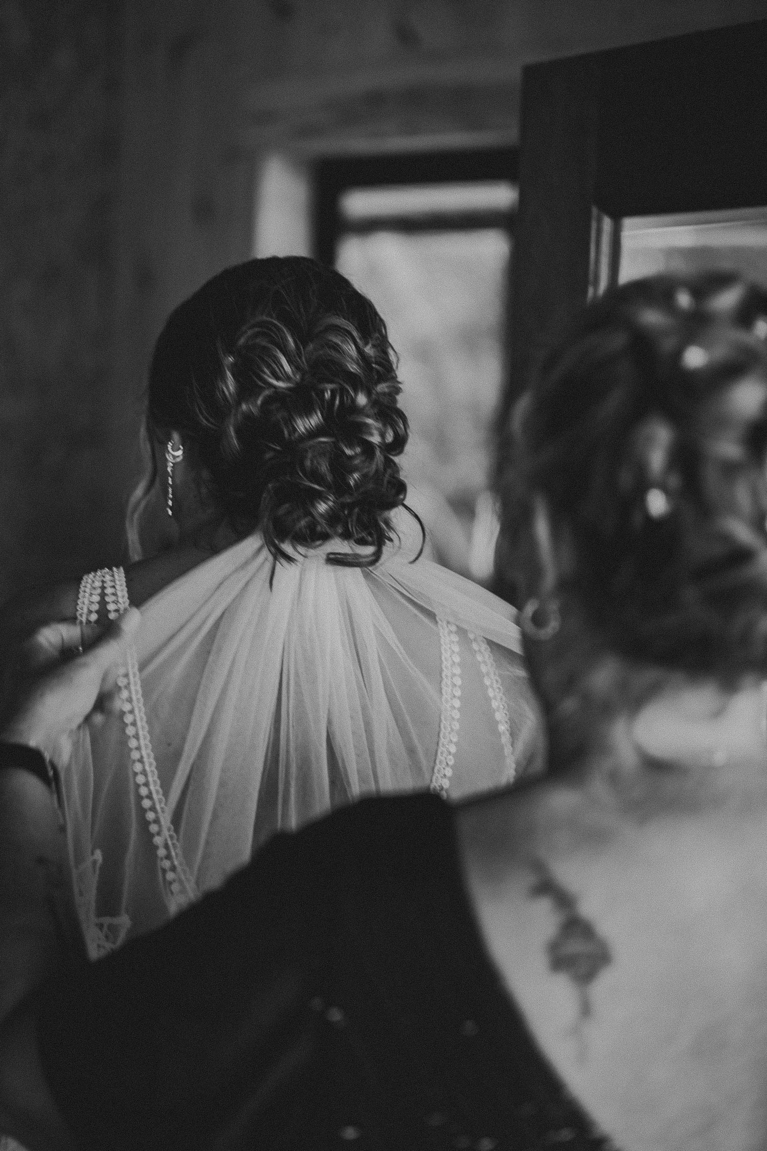 Black and white photo of bride's elegant updo with pearl-trimmed veil being adjusted by bridesmaid.