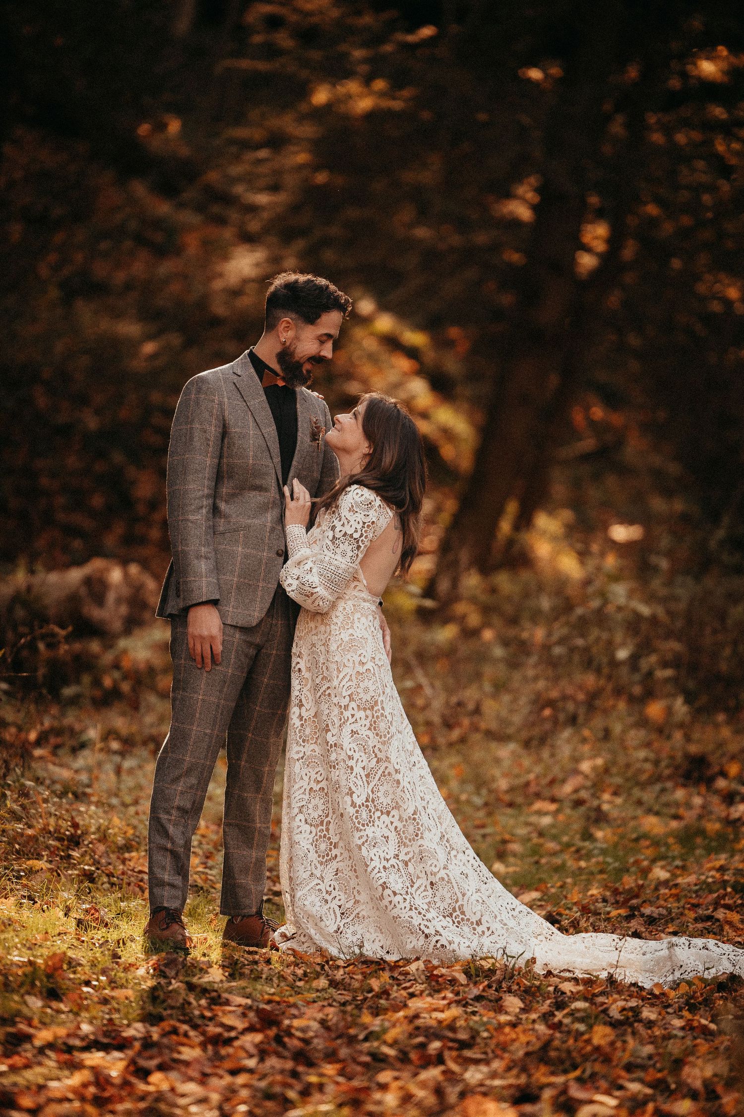 Bride and groom embrace romantically in autumn forest surrounded by golden fallen leaves.
