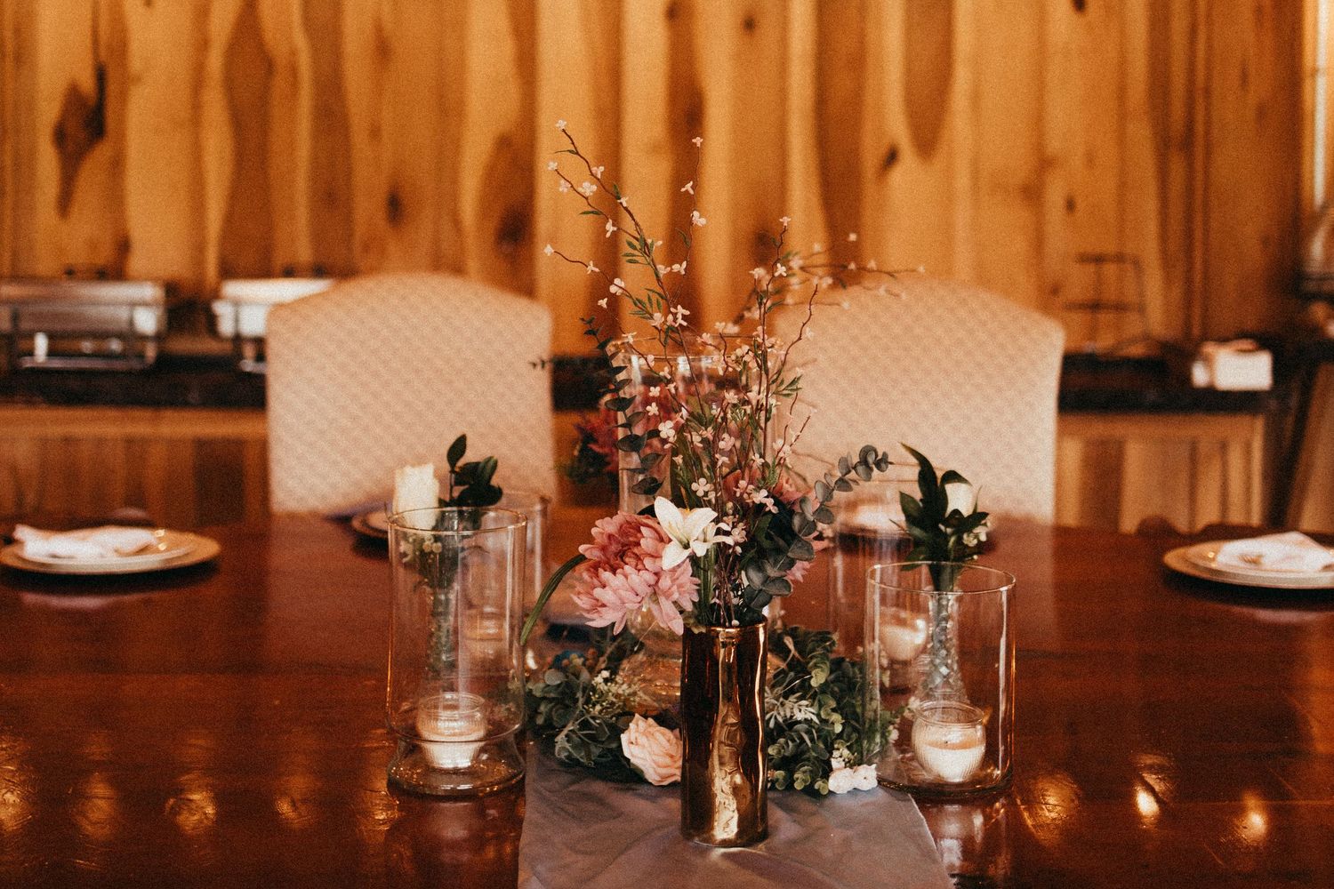 Elegant wedding reception table with floral centerpiece, candles, and pink blooms in wood-paneled venue.