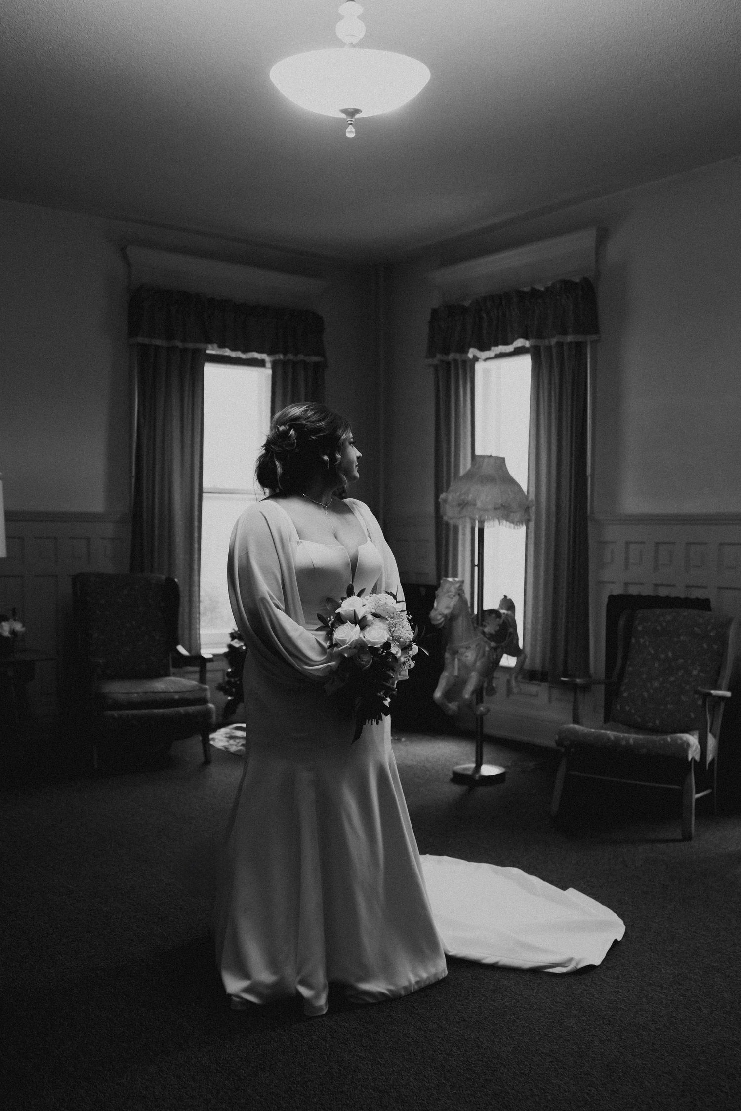 Black and white portrait of bride in elegant gown holding bouquet by vintage room windows.