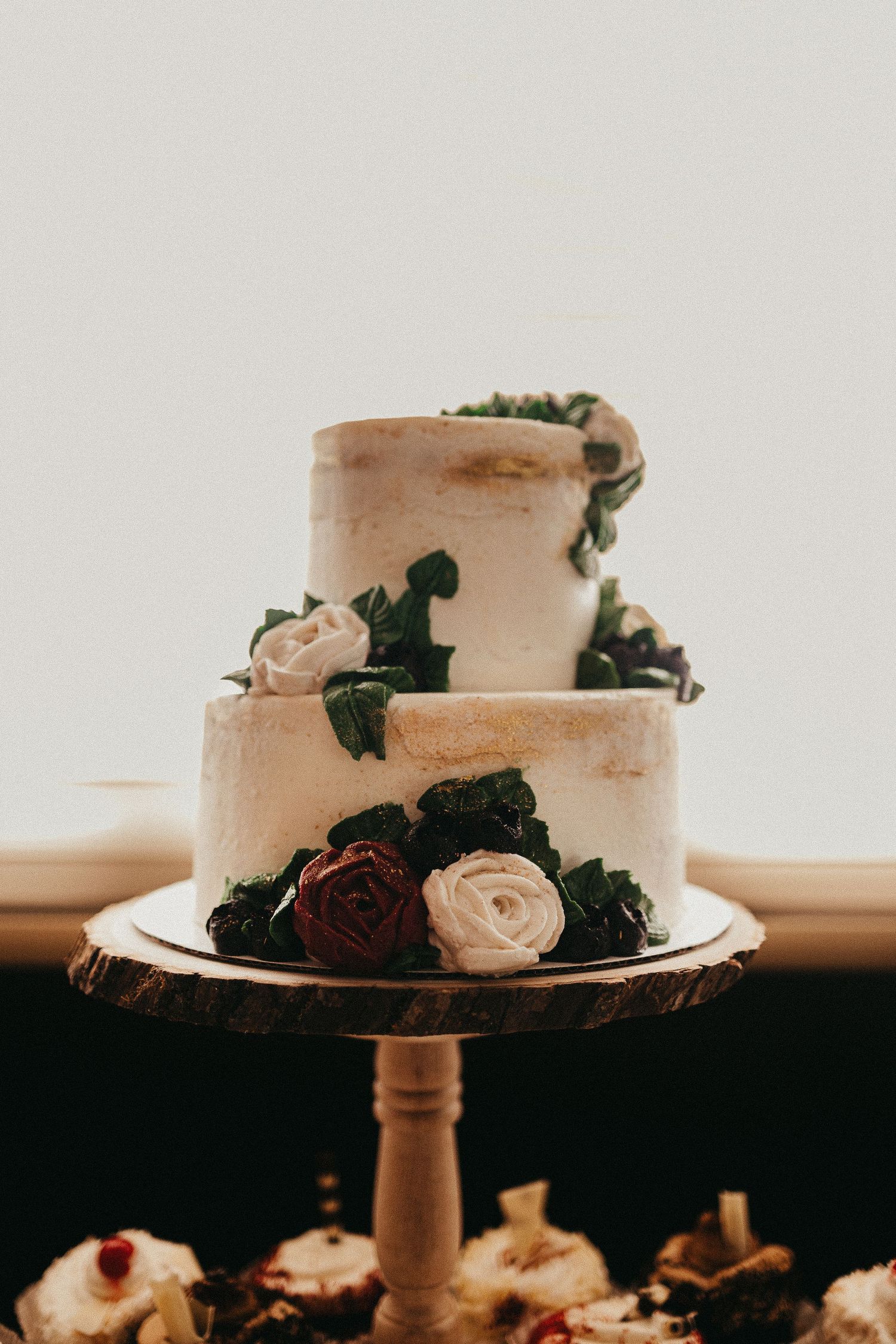 Two-tier semi-naked wedding cake adorned with white and burgundy roses and greenery on a rustic wood cake stand.