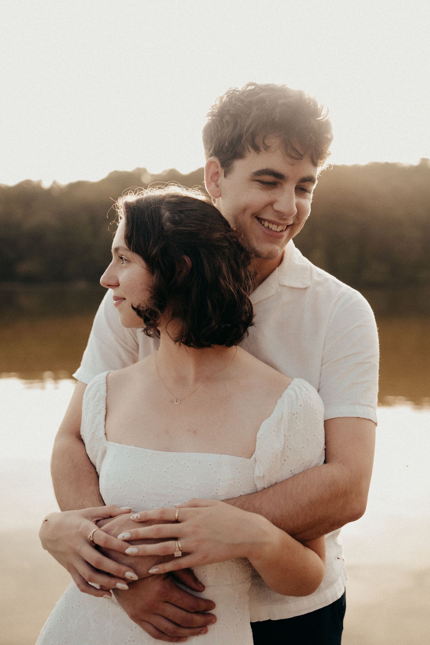 Smiling young couple embracing near a lake at sunset, man in white shirt holding woman in white dress.