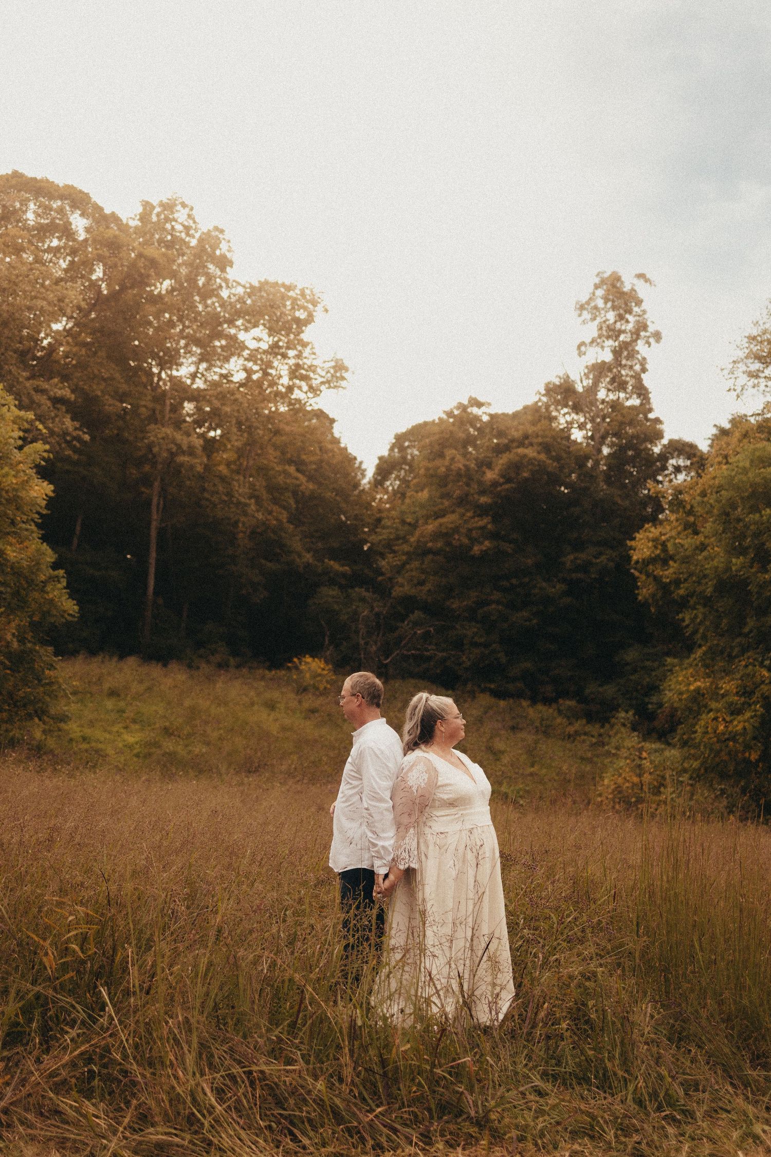 Bride and groom stand back to back holding hands in golden meadow surrounded by lush trees.