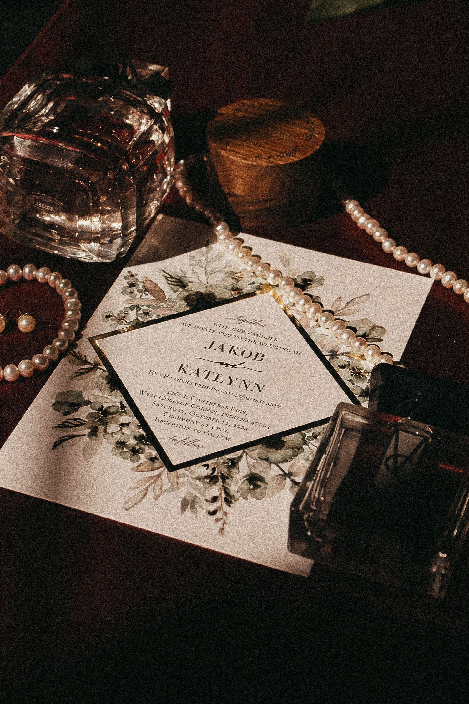 Elegant fall wedding invitation for Jakob and Katlynn surrounded by pearl necklaces and perfume bottles on a dark wood surface.