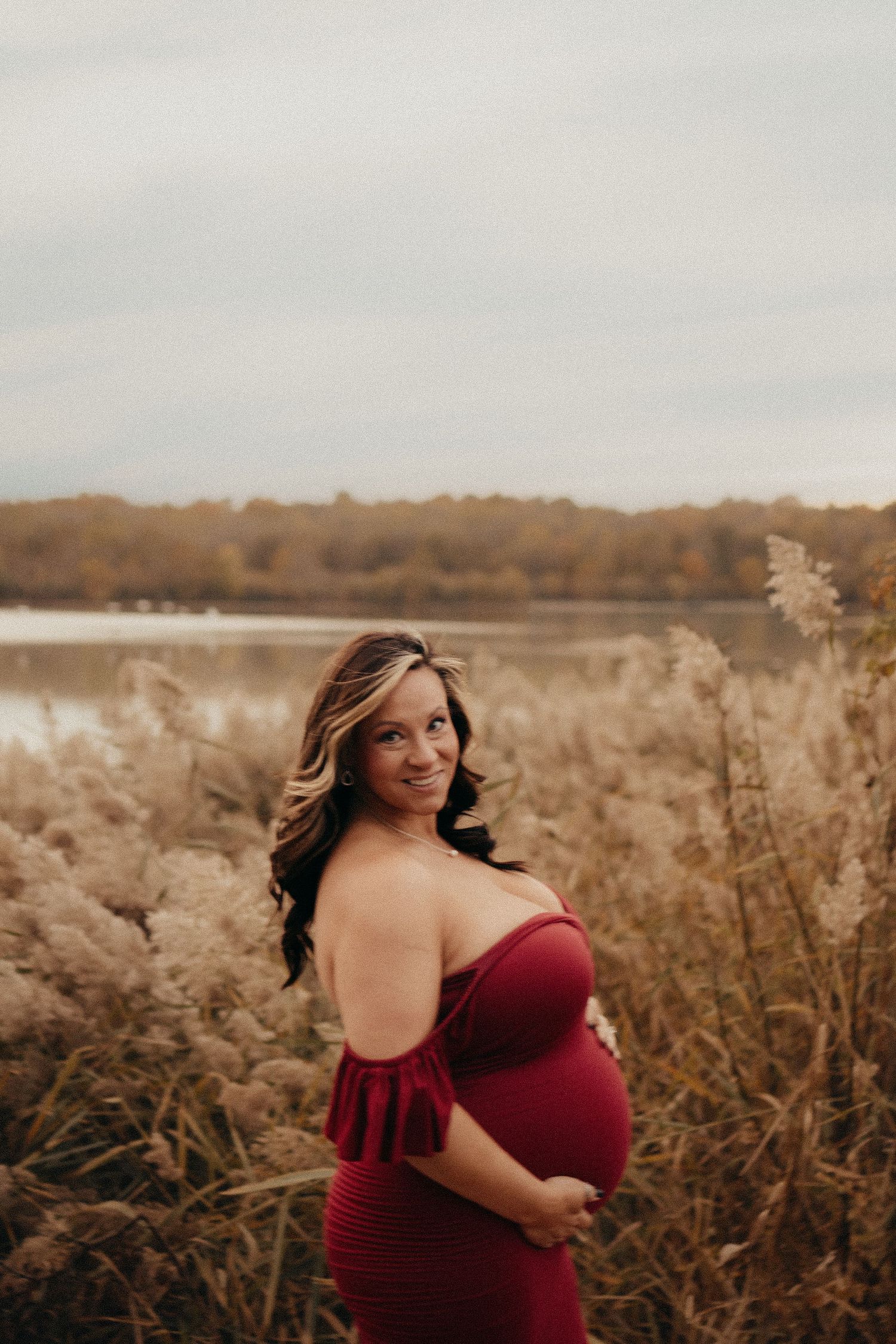 Pregnant woman in red off-shoulder dress posing outdoors near a lake surrounded by tall golden grasses.