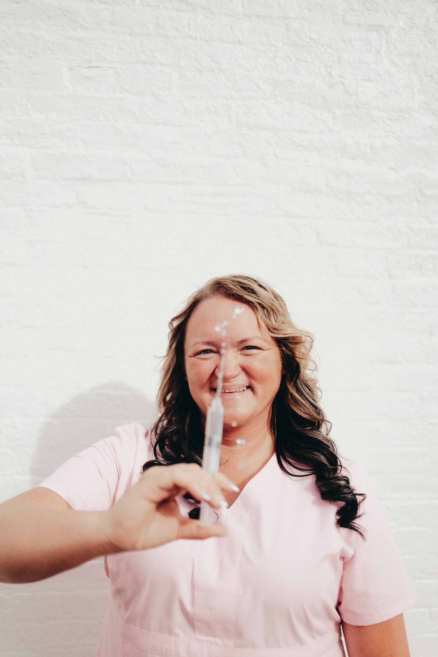 Smiling female nurse in pink scrubs holding a syringe toward the camera against a white wall.