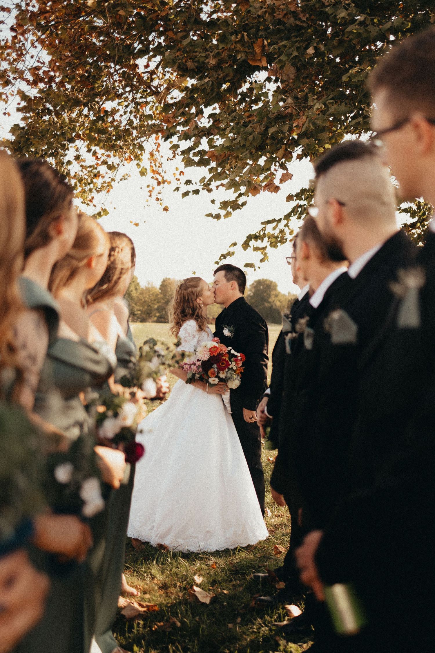 Bride and groom share a kiss surrounded by wedding party under autumn foliage in golden outdoor light.