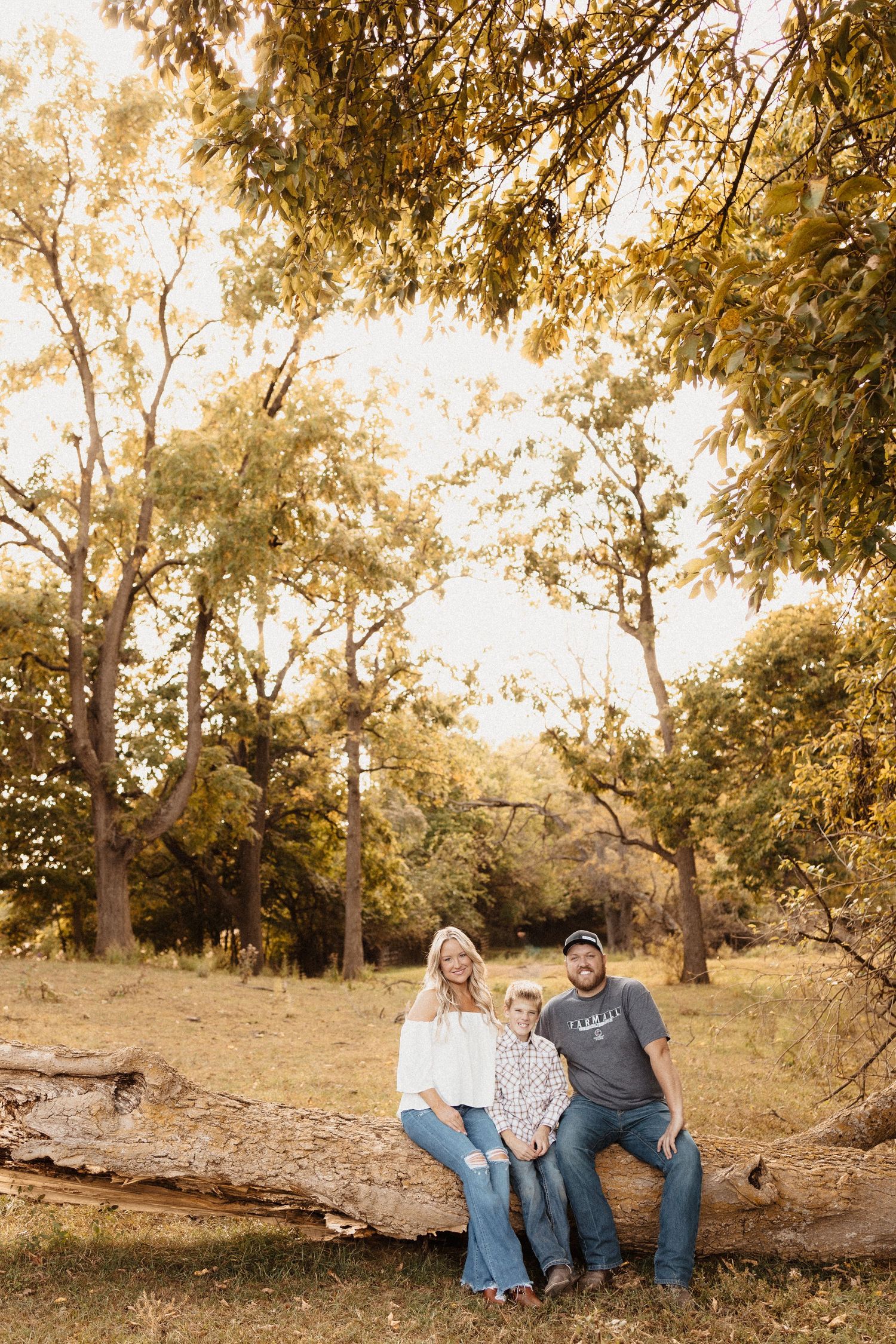 A happy family of three poses on a fallen log in a sunlit autumn forest setting.