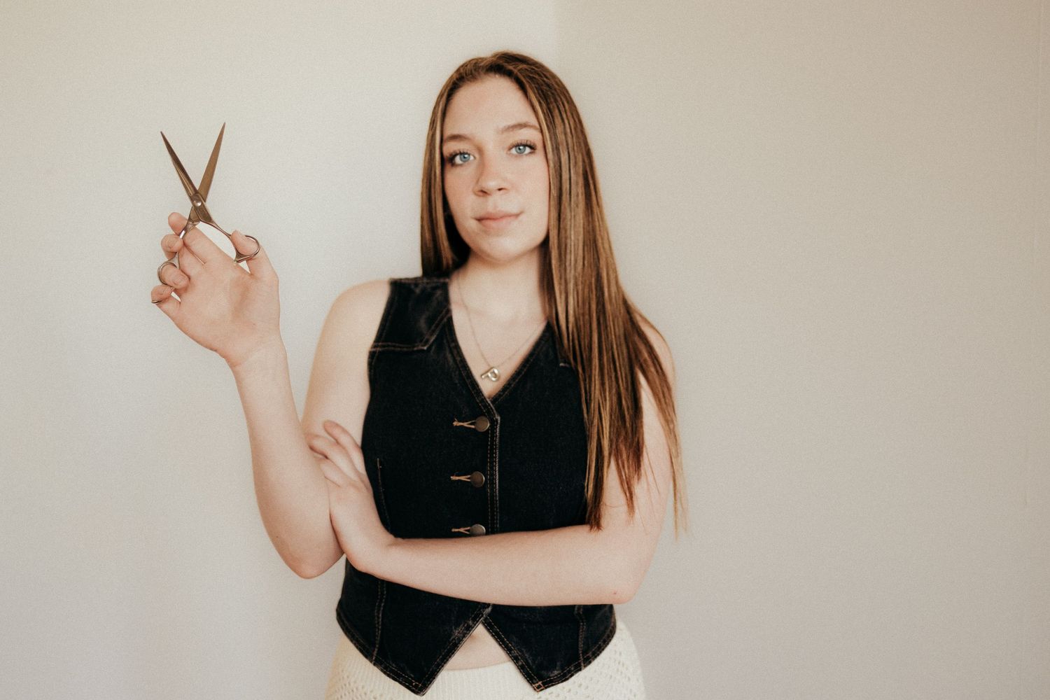 Young woman holding scissors with arms crossed against a neutral white wall.