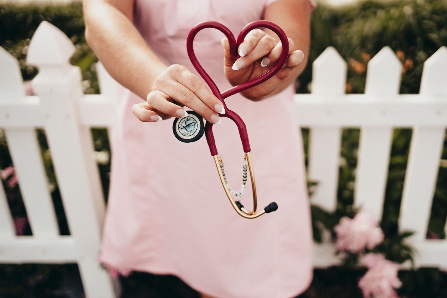 Nurse holding a red stethoscope shaped into a heart symbol outdoors near a white picket fence.