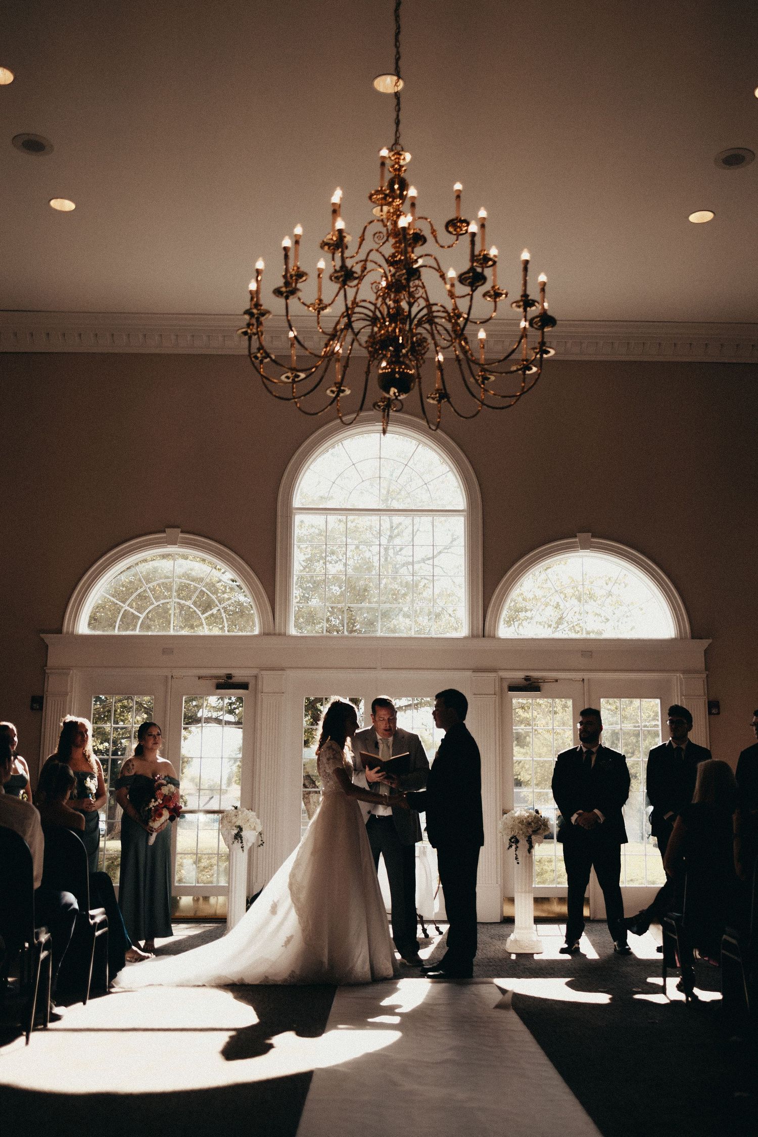 Bride and groom exchange vows beneath a grand chandelier in an elegant indoor wedding ceremony venue.
