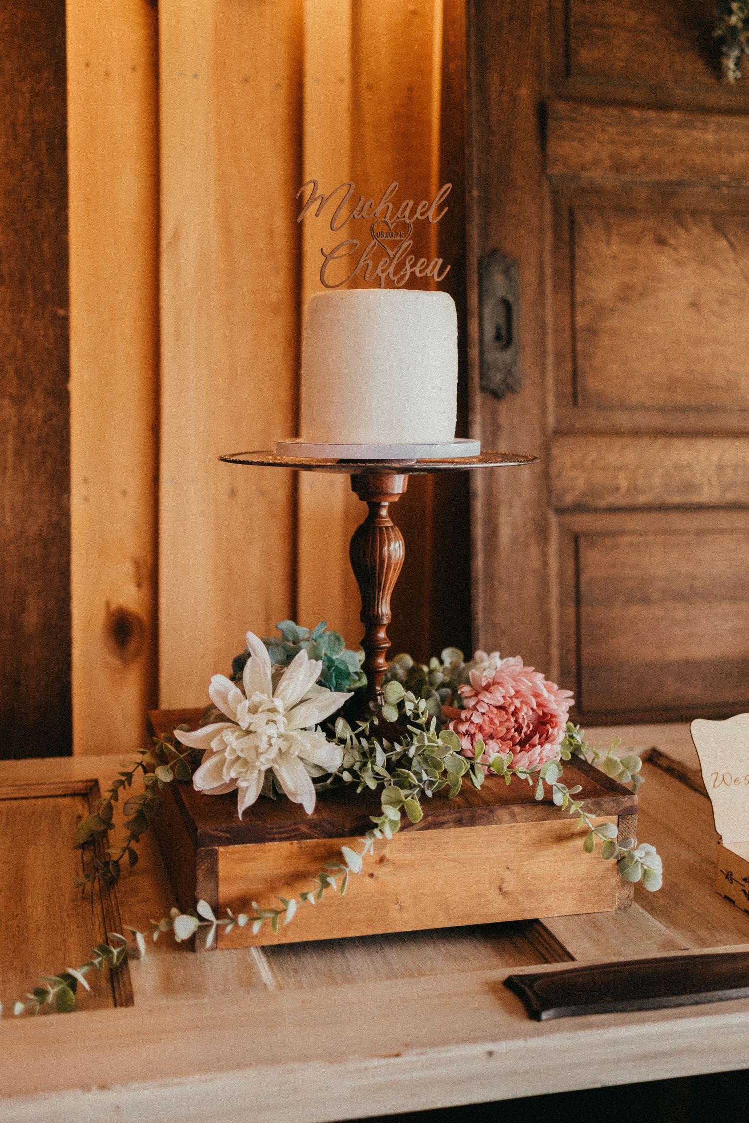Simple white wedding cake with Michael and Chelsea topper displayed on wooden pedestal with florals.